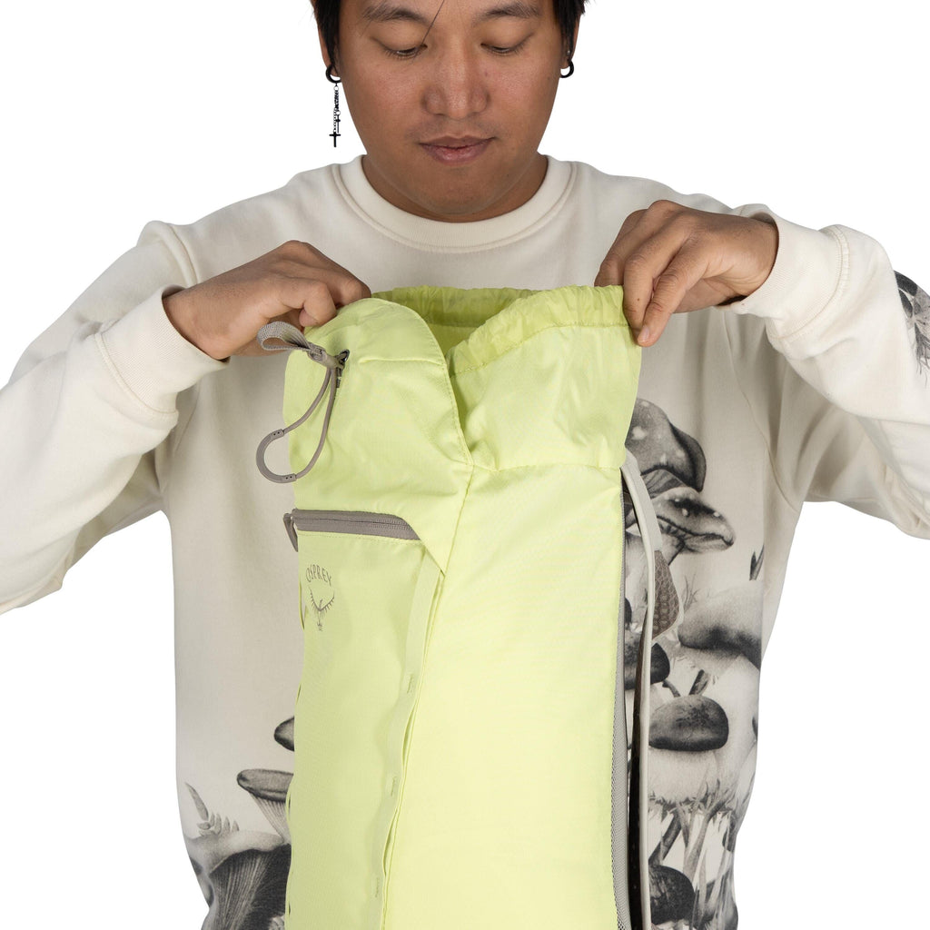The image shows a person holding open a light green backpack. The backpack features a simple drawstring closure and has a small front zippered pocket. The brand logo, "Osprey," is visible on the front of the backpack. The person is wearing a white sweatsh