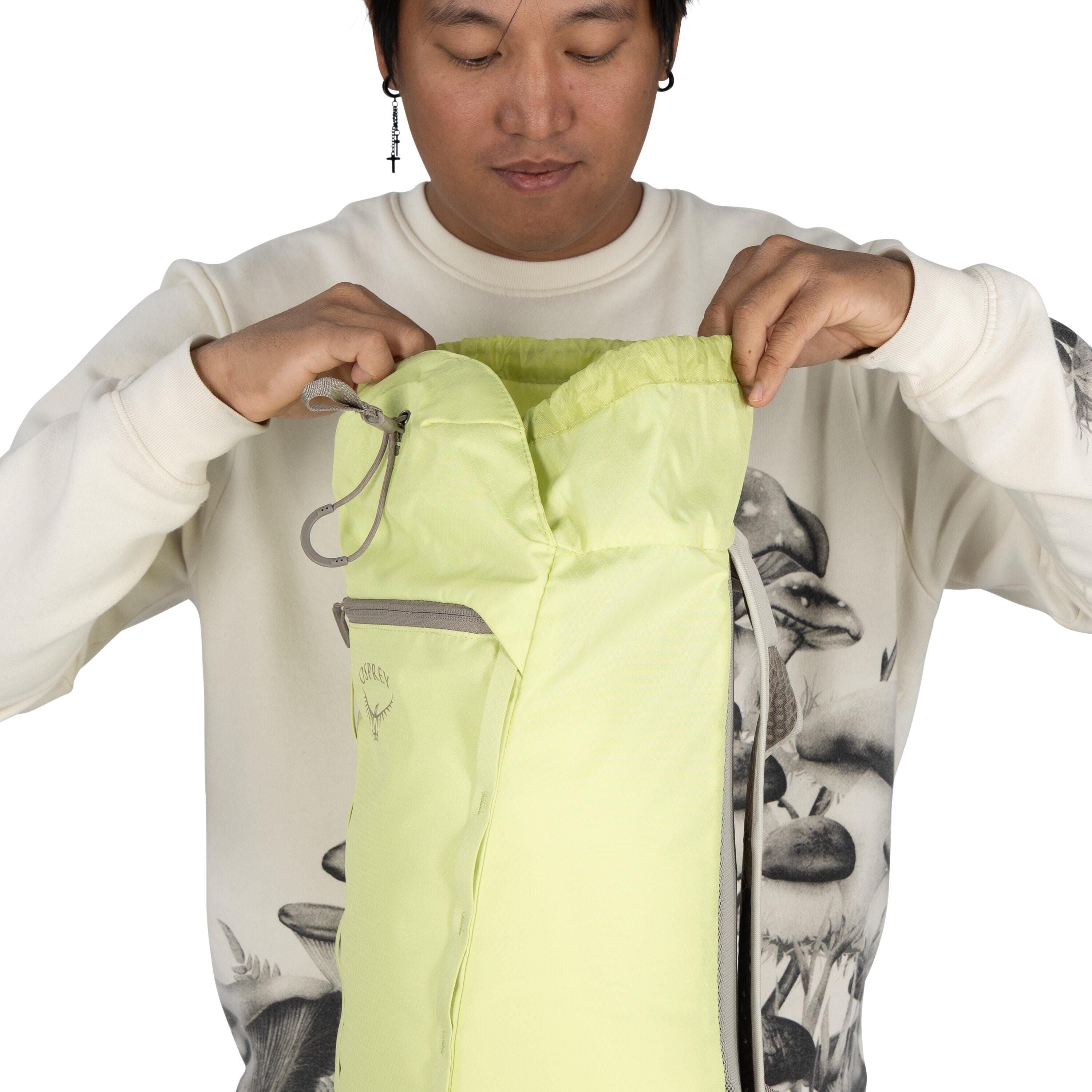 The image shows a person holding open a light green backpack. The backpack features a simple drawstring closure and has a small front zippered pocket. The brand logo, "Osprey," is visible on the front of the backpack. The person is wearing a white sweatsh