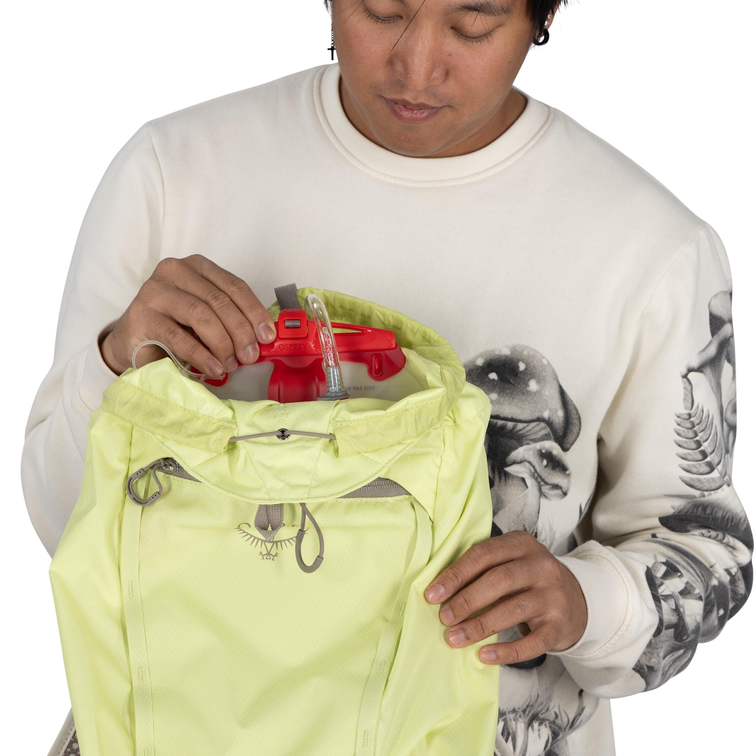 The image shows a person holding a light green Osprey backpack. The individual is placing or removing a red hydration reservoir from the top compartment of the backpack. They are wearing a white sweatshirt with intricate designs, including mushrooms and f