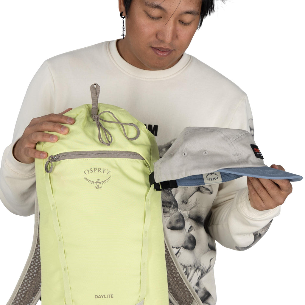 The image shows a person holding a light green backpack with the brand name "Osprey" on it, specifically labeled as the "Daylite" model. The backpack has a front zipper pocket and appears to be made of a lightweight material. In the other hand, the person