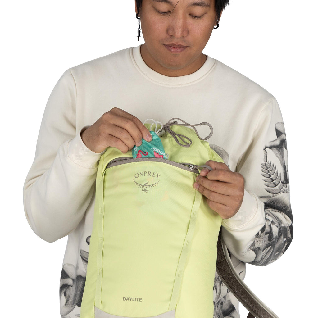 The image shows a person wearing a light-colored sweater with an artistic, grayscale print on the sleeves and torso. They are holding a light green Osprey Daylite backpack, opening its front zipper pocket. Inside the pocket, there is a colorful item with