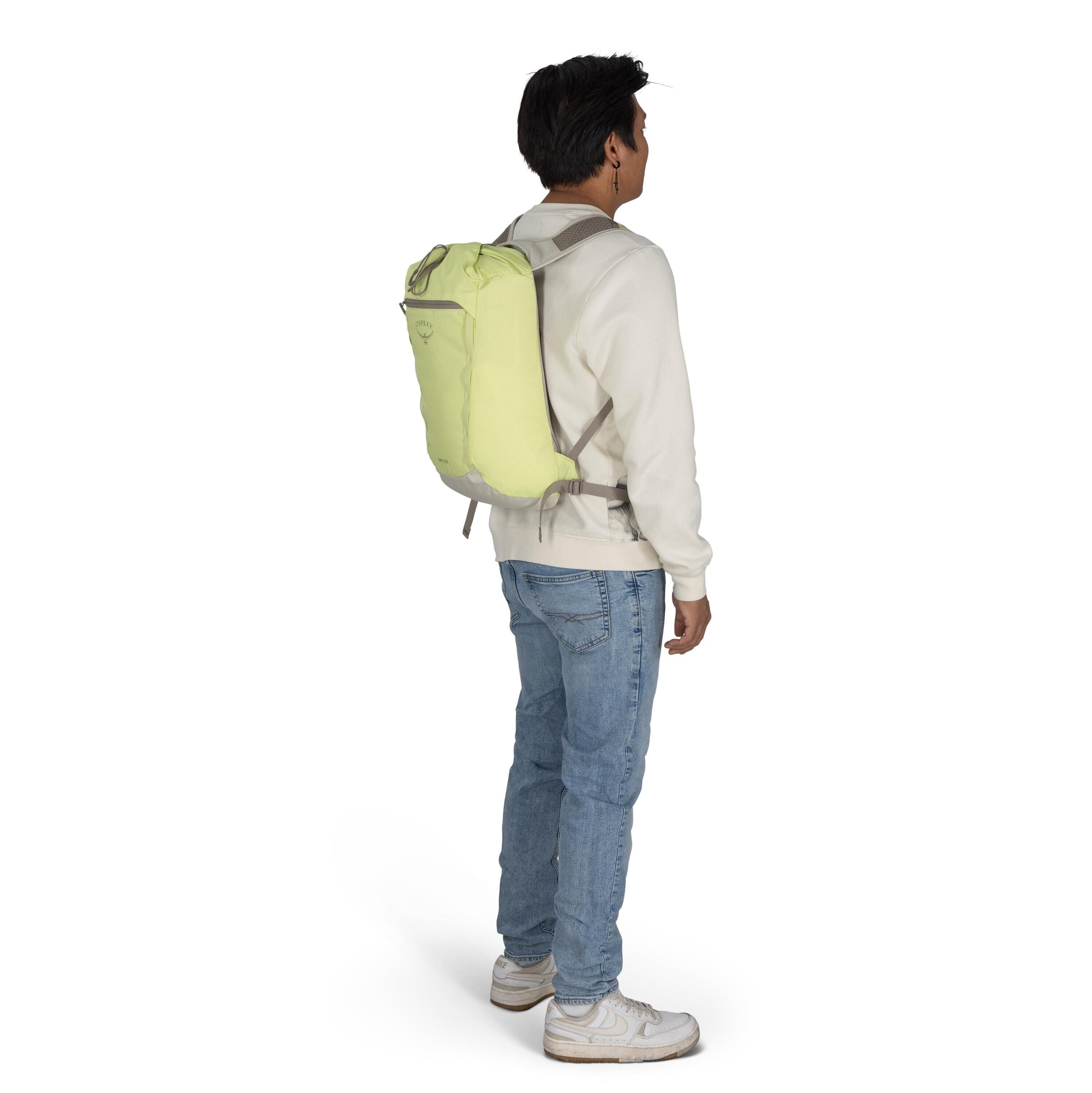 The image features a person standing sideways, wearing a light-colored sweatshirt and light blue jeans. They have on white sneakers and are carrying a light green backpack with grey straps. The backpack has the "Osprey" logo and the model name "Ultralight