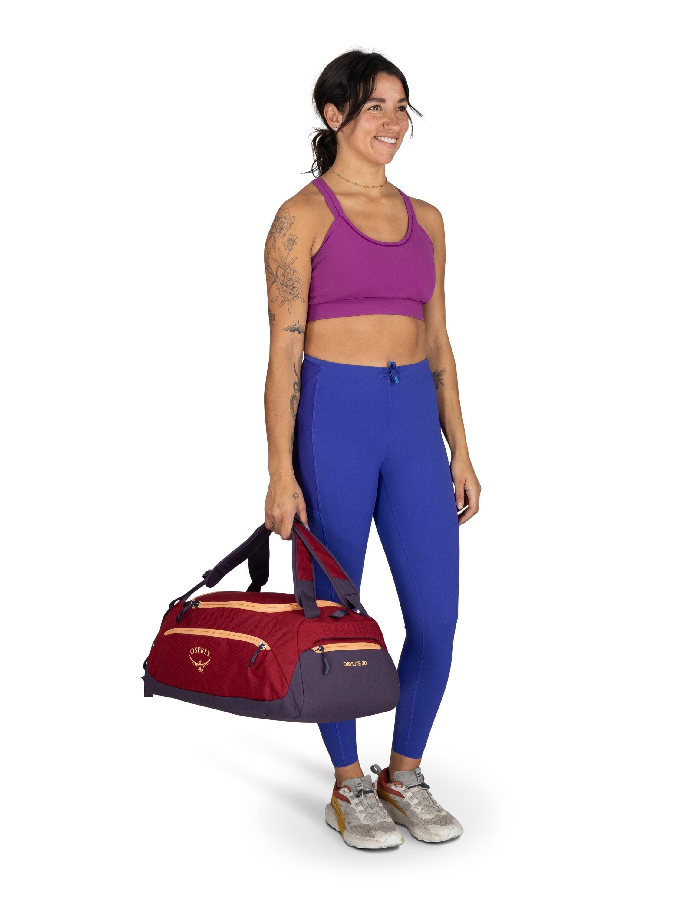 The image shows a person standing against a plain background, dressed in athletic attire. They are wearing a purple sports bra and blue leggings, paired with gray sneakers that have red accents. The person is holding a red and purple gym bag in their righ