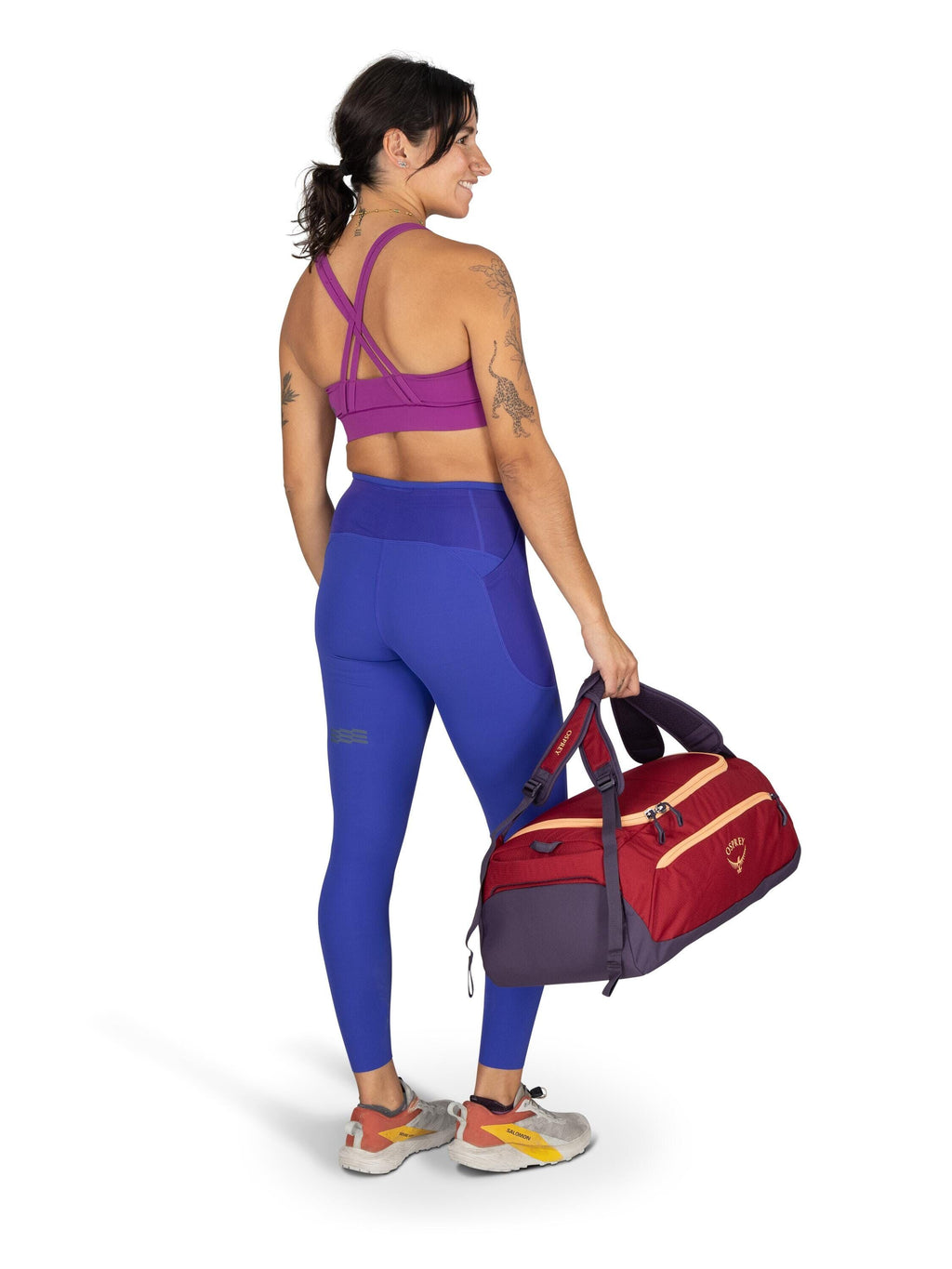The image shows a woman standing and facing away from the camera, dressed in athletic wear. She is wearing a magenta sports bra with cross-back straps, and vivid blue high-waisted leggings that have pocket details on the sides. The woman has several tatto