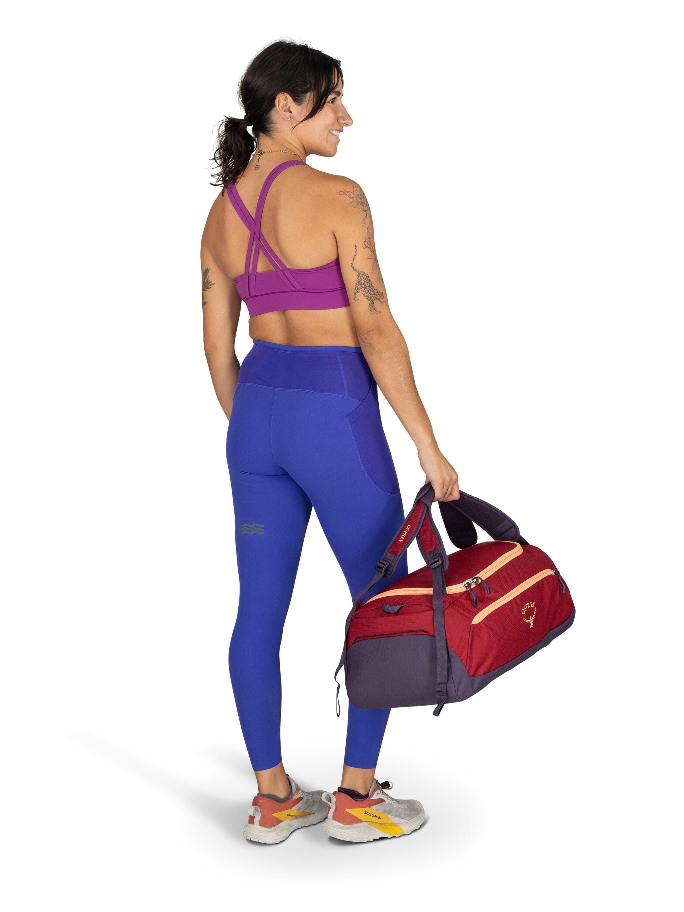 The image shows a woman standing and facing away from the camera, dressed in athletic wear. She is wearing a magenta sports bra with cross-back straps, and vivid blue high-waisted leggings that have pocket details on the sides. The woman has several tatto