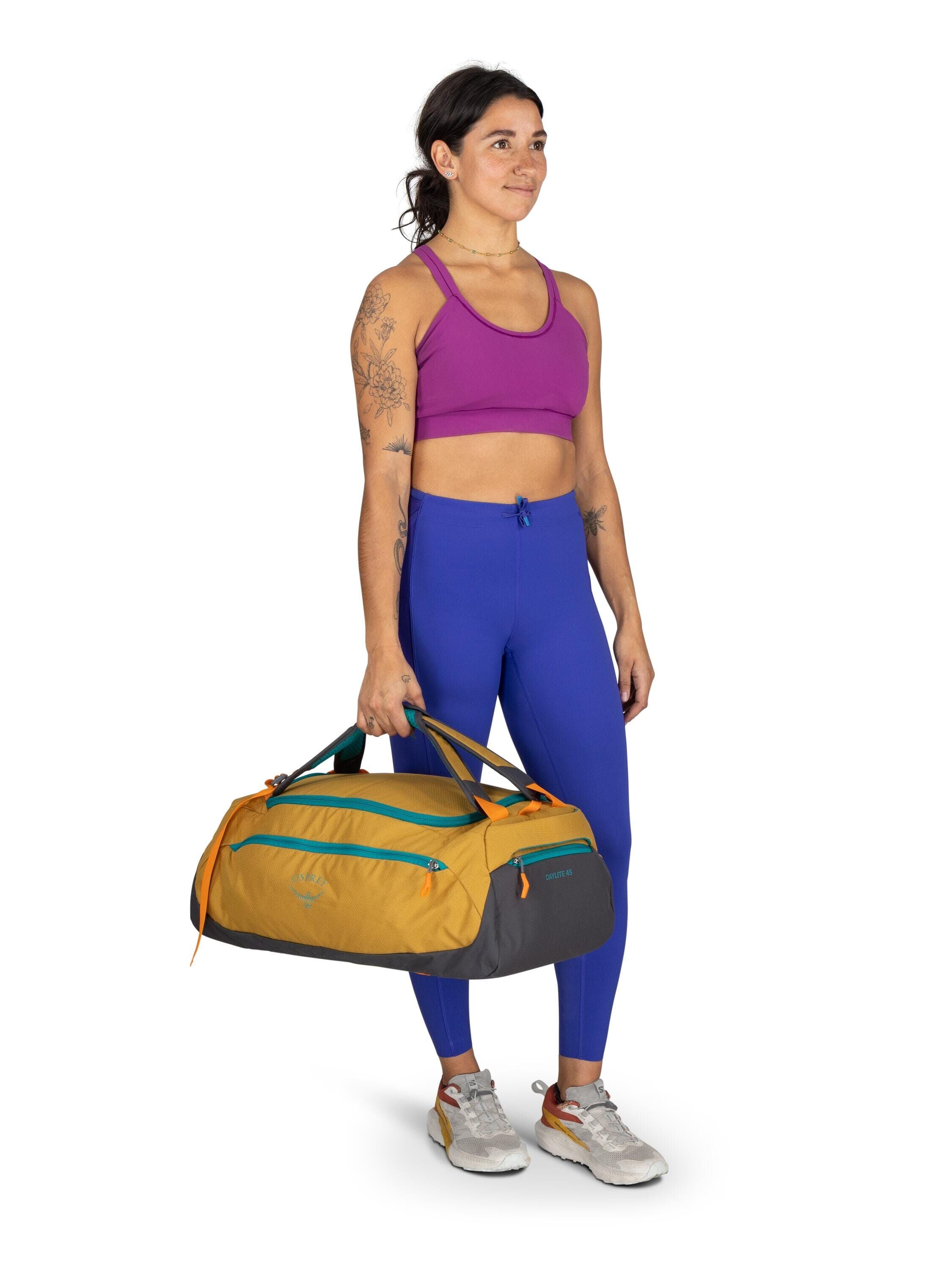 The image shows a person standing and holding a large, stylish duffle bag. The individual is wearing athletic attire, including a vibrant purple sports bra and blue leggings, paired with light-colored sneakers. The duffle bag is prominently designed with