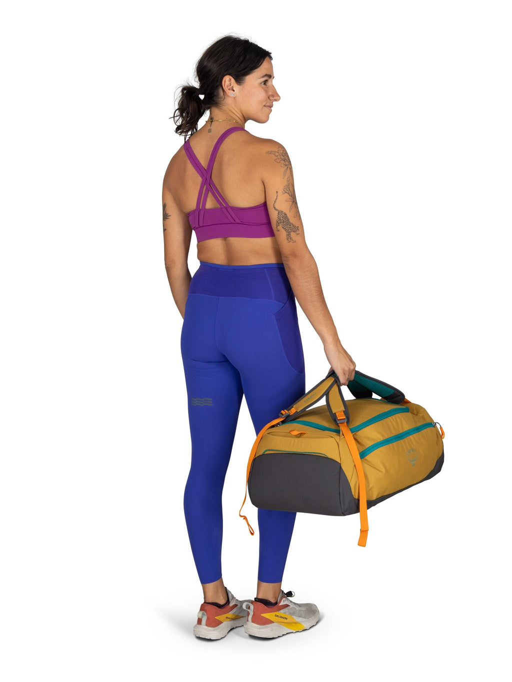 The image features a woman standing and facing away from the camera. She is wearing a vibrant sports outfit, which includes a purple sports bra and blue leggings with a stripe detail. Her hair is tied back in a ponytail, and she has visible tattoos on her