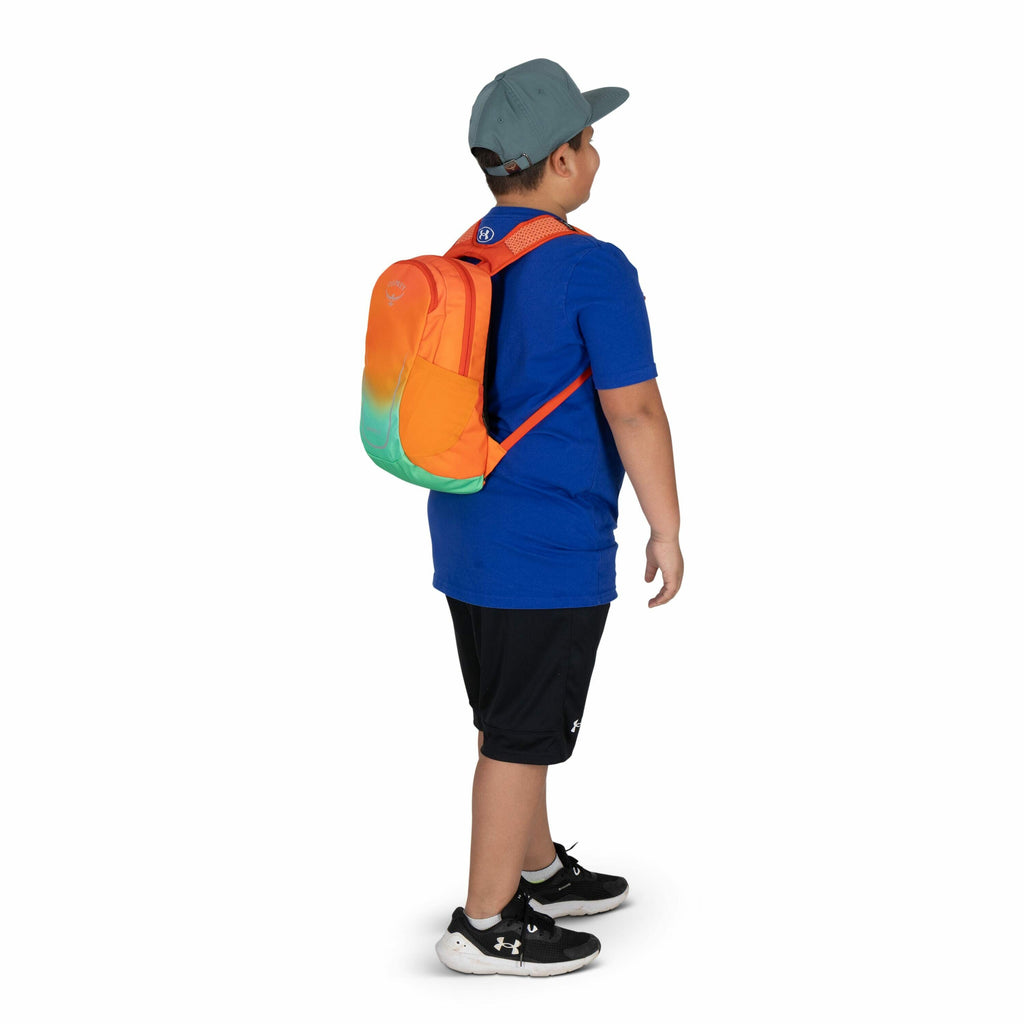 The image features a young boy standing and facing slightly to the right, showcasing a colorful backpack he is wearing. The backpack has a vibrant design with a gradient of orange transitioning to green at the bottom. The boy is dressed casually, wearing