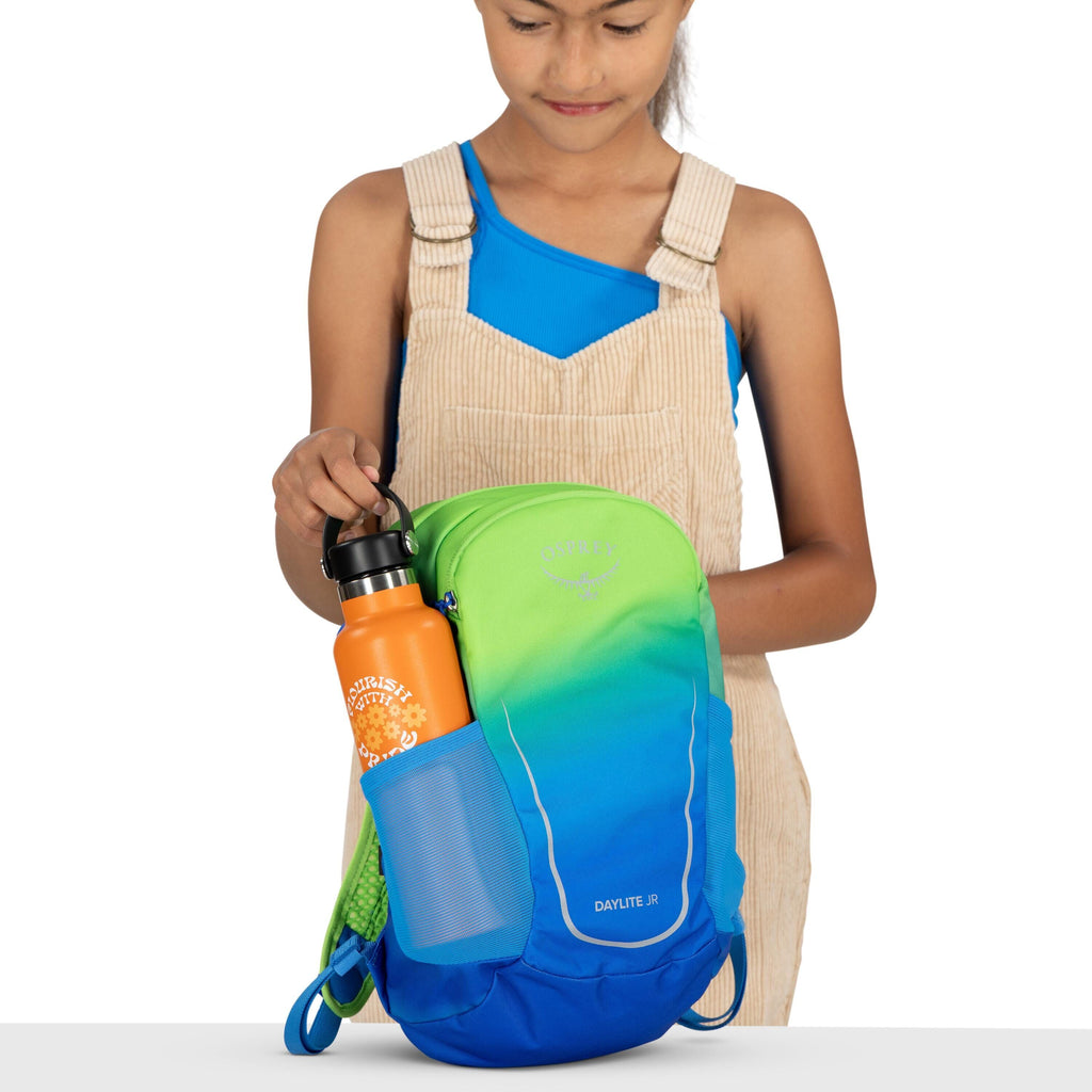 The image shows a person holding a colorful backpack with their right hand. The backpack is an Osprey Daylite Jr, featuring a gradient design that transitions from bright green at the top to deep blue at the bottom. The person is wearing a blue sleeveless