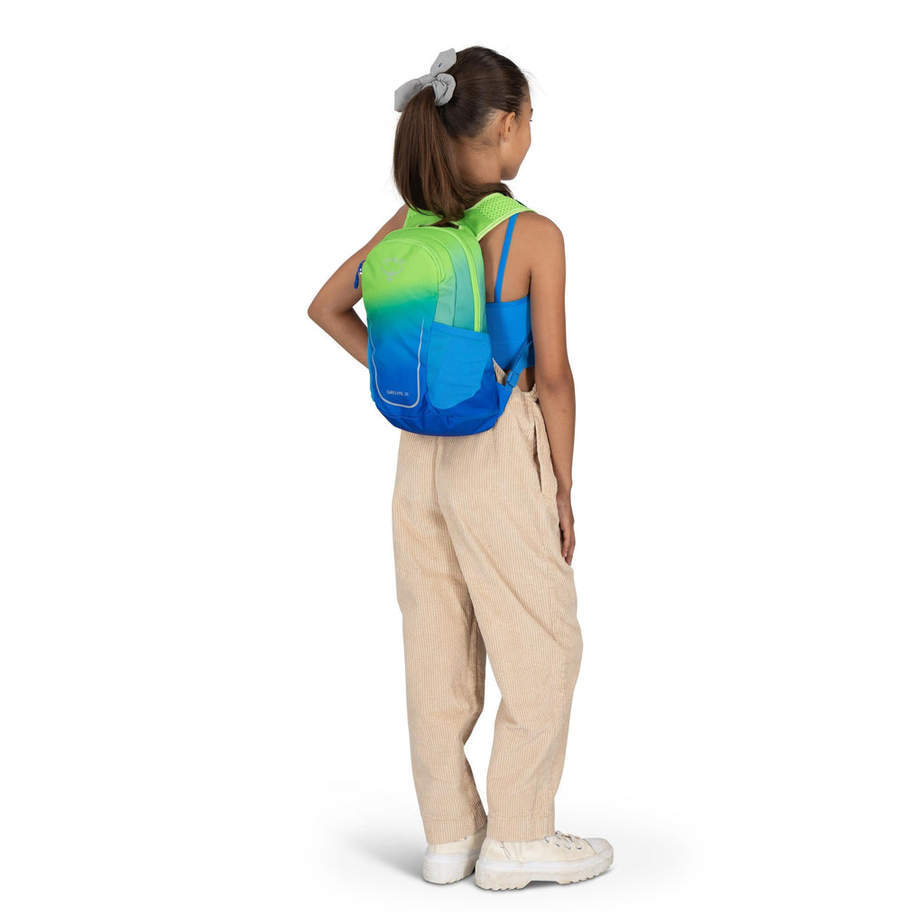 The image features a young girl standing in profile, looking to her right. She is wearing a vibrant backpack with a gradient design that transitions from green at the top to blue at the bottom. Her outfit includes a blue tank top and beige, loose-fitting