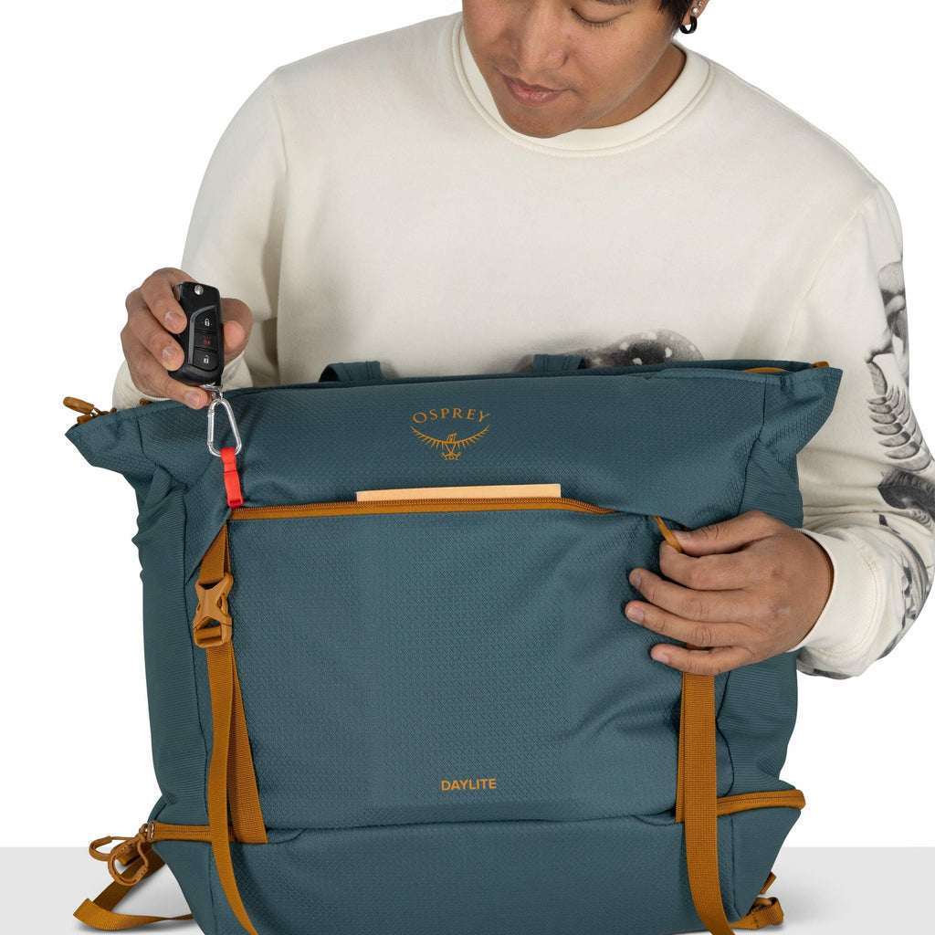 The image depicts a person wearing a white sweater with dark artistic designs on the sleeves, holding a teal-colored Osprey backpack. The backpack has distinct tan accents, including straps and zippers. The person is using a carabiner attached to the bag'