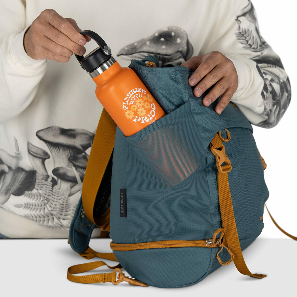 The image shows a person wearing a sweatshirt with a mushroom design, placing an orange water bottle into a side pocket of a teal backpack. The bottle is labeled "FLOURISH WITH PRIDE" and features flowers in its design. The backpack has mustard yellow str