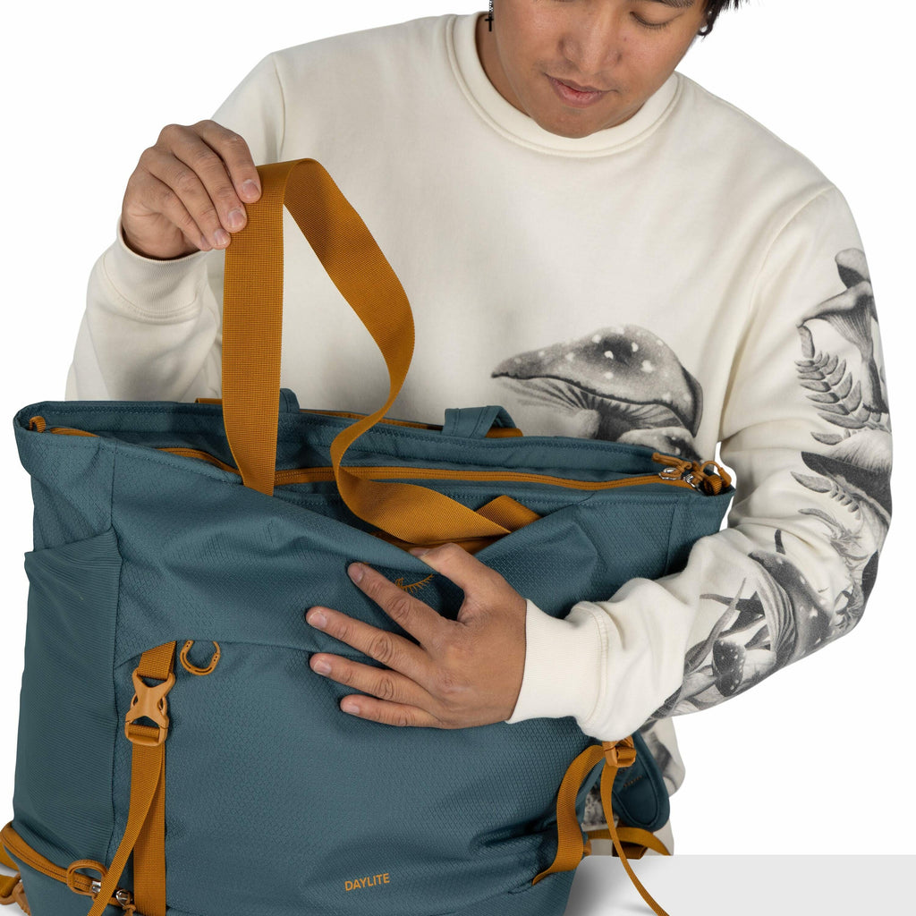 The image shows a person holding a stylish teal-colored bag with mustard yellow straps and accents. The bag has the word "DAYLITE" written on it, indicating it might be a product of the Osprey Daylite series. The person is wearing a light-colored sweatshi