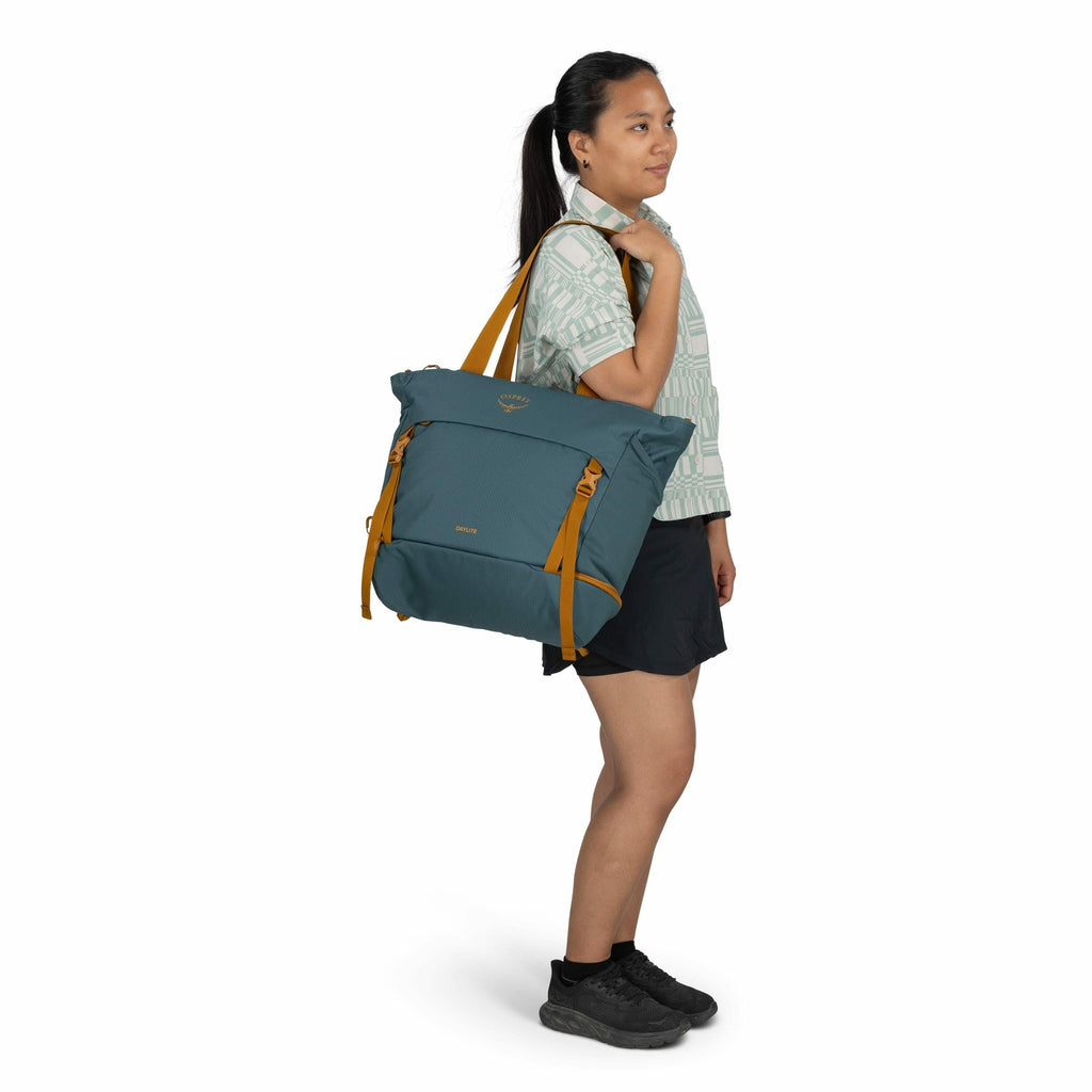 The image shows a person dressed casually, carrying a large Osprey-branded backpack or tote. The bag is a shade of teal blue with mustard-yellow straps and features the logo and the word "Arcane" prominently on the front. The individual is wearing a patte