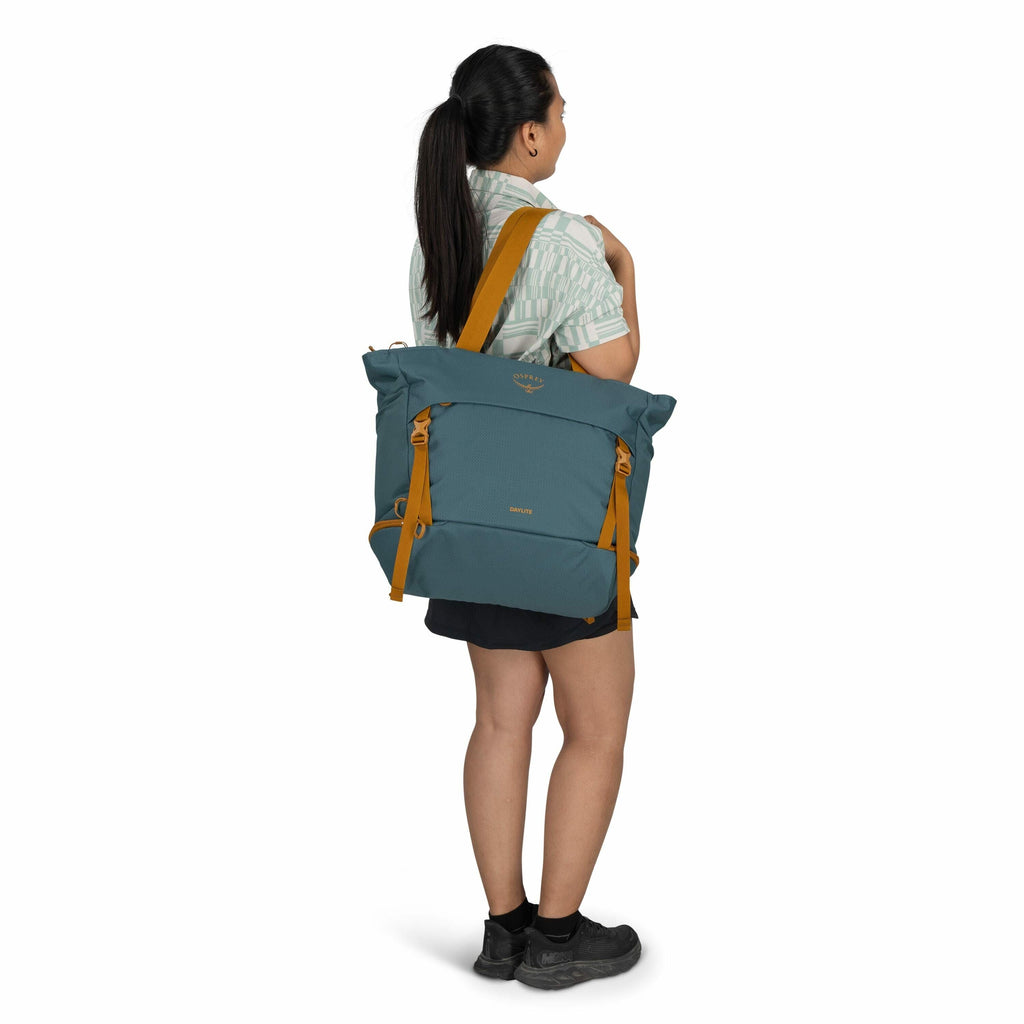 The image shows a woman with long, dark hair tied in a ponytail, standing and facing slightly away from the camera. She is wearing a light-colored patterned shirt, black shorts, and black shoes. Over her shoulder, she is carrying a large teal Osprey tote