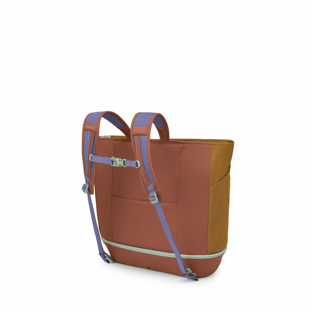 The image shows a colorful backpack from a rear angle, highlighting its design and functionality. The backpack features a combination of earthy brown and mustard colors for its main body, with lilac-colored shoulder straps that include light blue padding