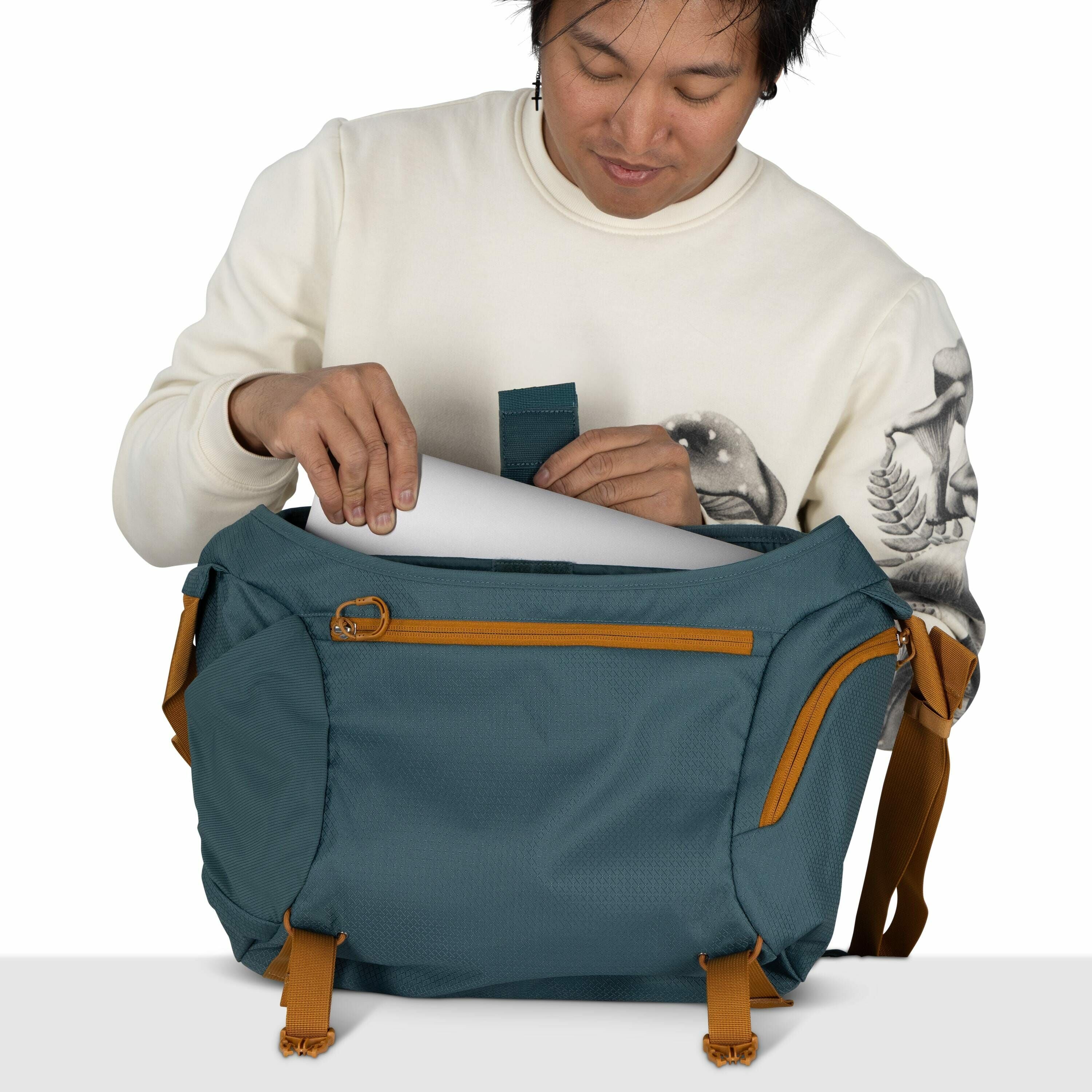 The image depicts a person holding a teal and mustard-yellow colored duffel bag. The person is wearing a cream-colored sweatshirt with artistic designs on the sleeves, featuring mushrooms and trees. They are putting a white sheet of paper into the main co