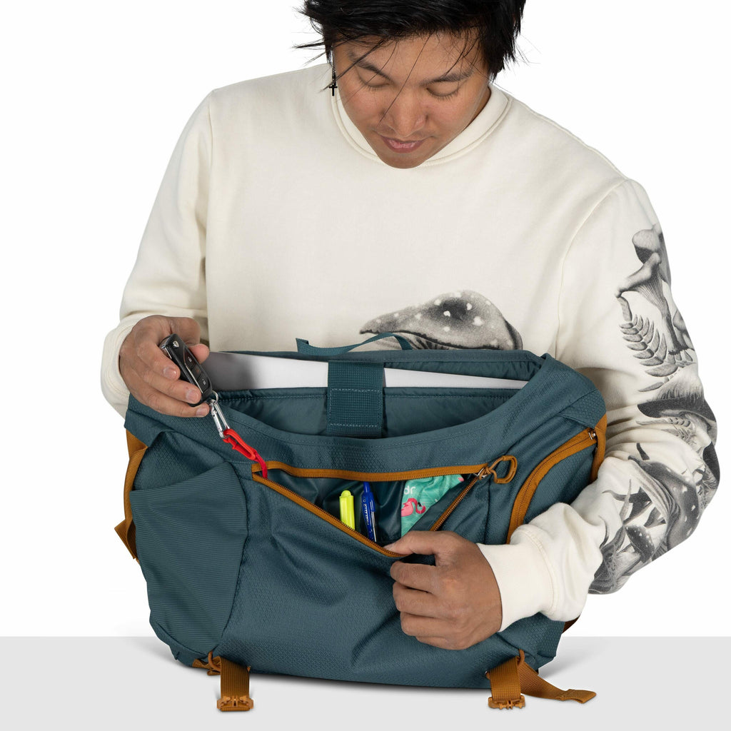 The image shows a person wearing a cream-colored sweatshirt with a detailed bird design on the sleeves and a mushroom illustration on the chest. They are holding a teal-colored bag with mustard-colored straps and zipper linings. The bag's main compartment