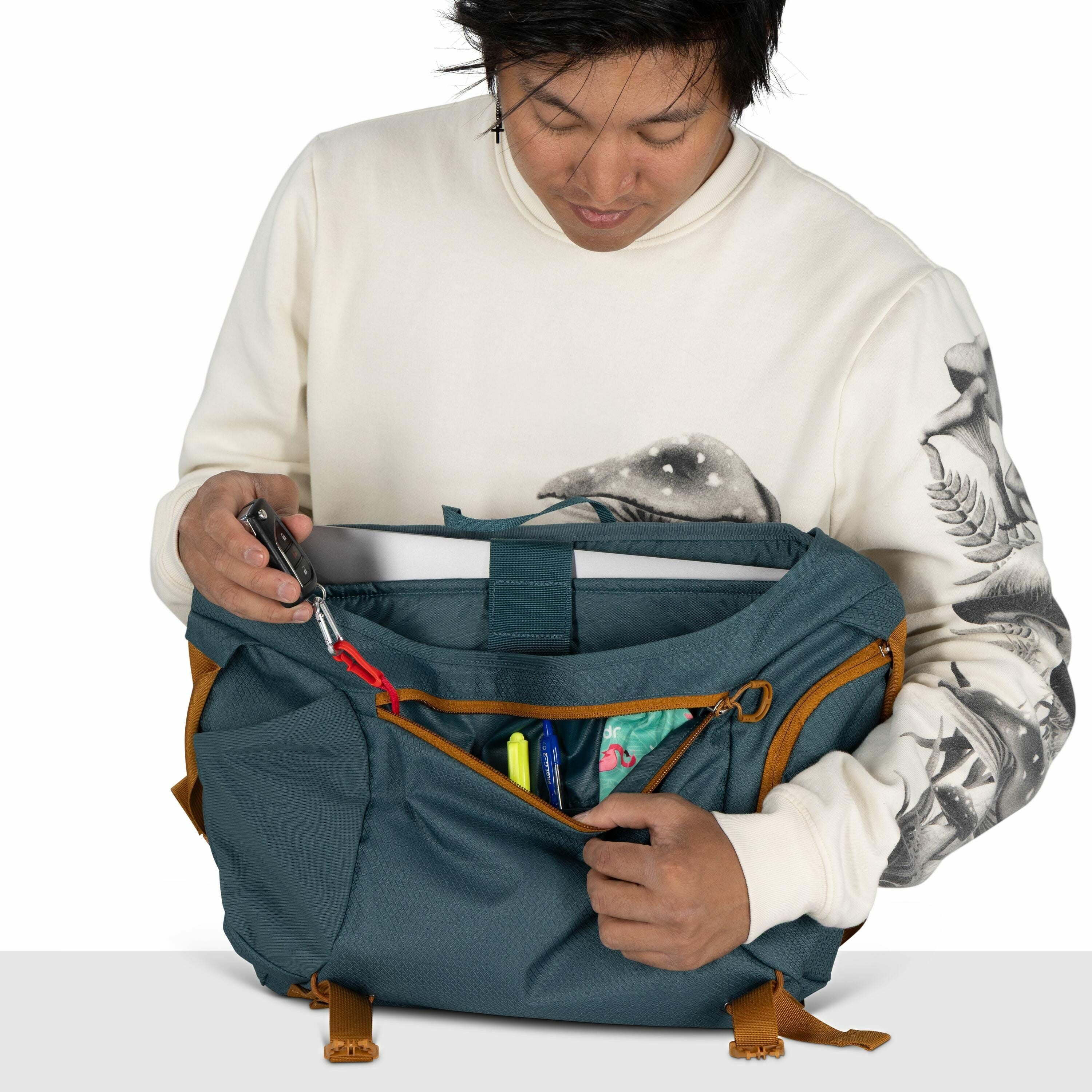 The image shows a person wearing a cream-colored sweatshirt with a detailed bird design on the sleeves and a mushroom illustration on the chest. They are holding a teal-colored bag with mustard-colored straps and zipper linings. The bag's main compartment