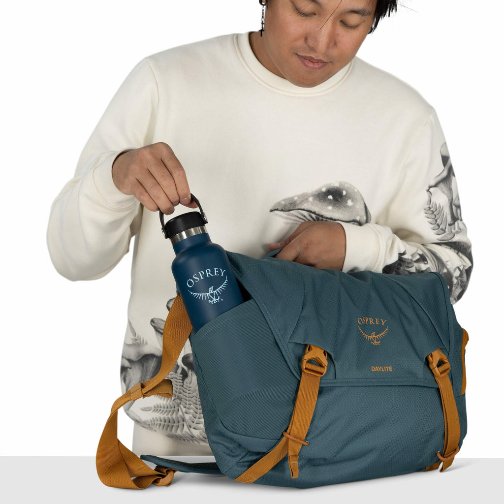 The image depicts a person holding a teal-colored Osprey Daylite bag with mustard-colored straps. The person is placing a blue Osprey-branded water bottle into one of the bag's pockets. The individual is wearing a cream-colored sweatshirt featuring a gray
