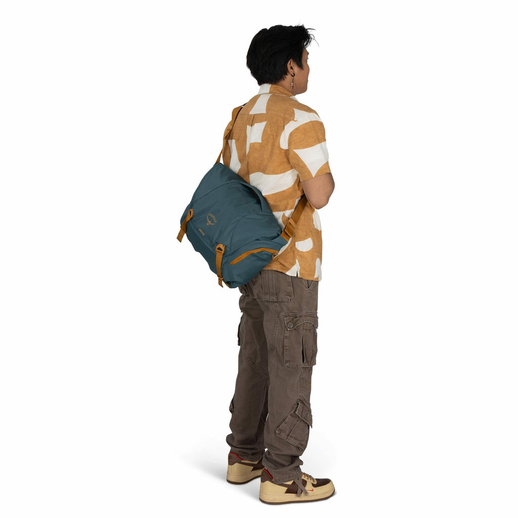 The image features a person standing and facing away, wearing a short-sleeved, button-up shirt with a bold orange and white pattern. They have a blue bag slung over their shoulder, featuring tan accents and the logo "Osprey." The person is dressed in brow