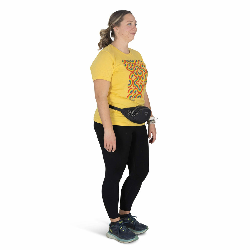 The image shows a woman standing and facing sideways, wearing a vibrant yellow t-shirt featuring an abstract, colorful pattern on the front. She pairs the t-shirt with black leggings and sporty sneakers in shades of blue, green, and yellow. Her outfit is