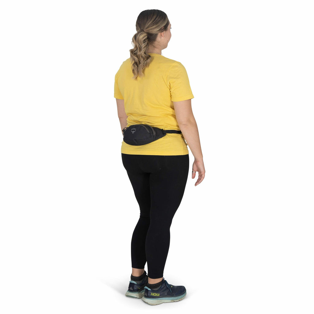 The image shows a woman standing and facing away from the camera. She is wearing a bright yellow t-shirt and black leggings, along with teal and black athletic shoes. Her hair is styled in a loose braid that falls over her shoulder. Around her waist, she