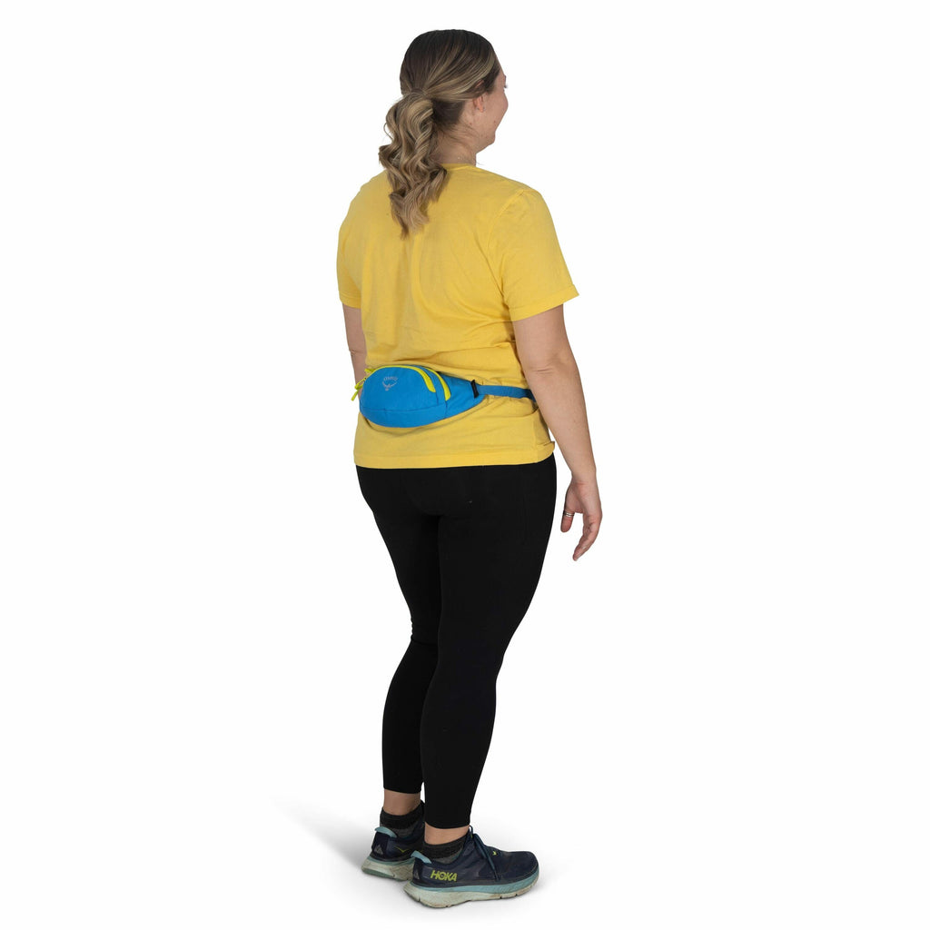 The image shows a woman standing and facing away from the camera. She is wearing a yellow T-shirt, black leggings, and dark athletic shoes with a distinctive green sole. Her hair is styled in a loose braid that rests over her shoulder. She is also wearing