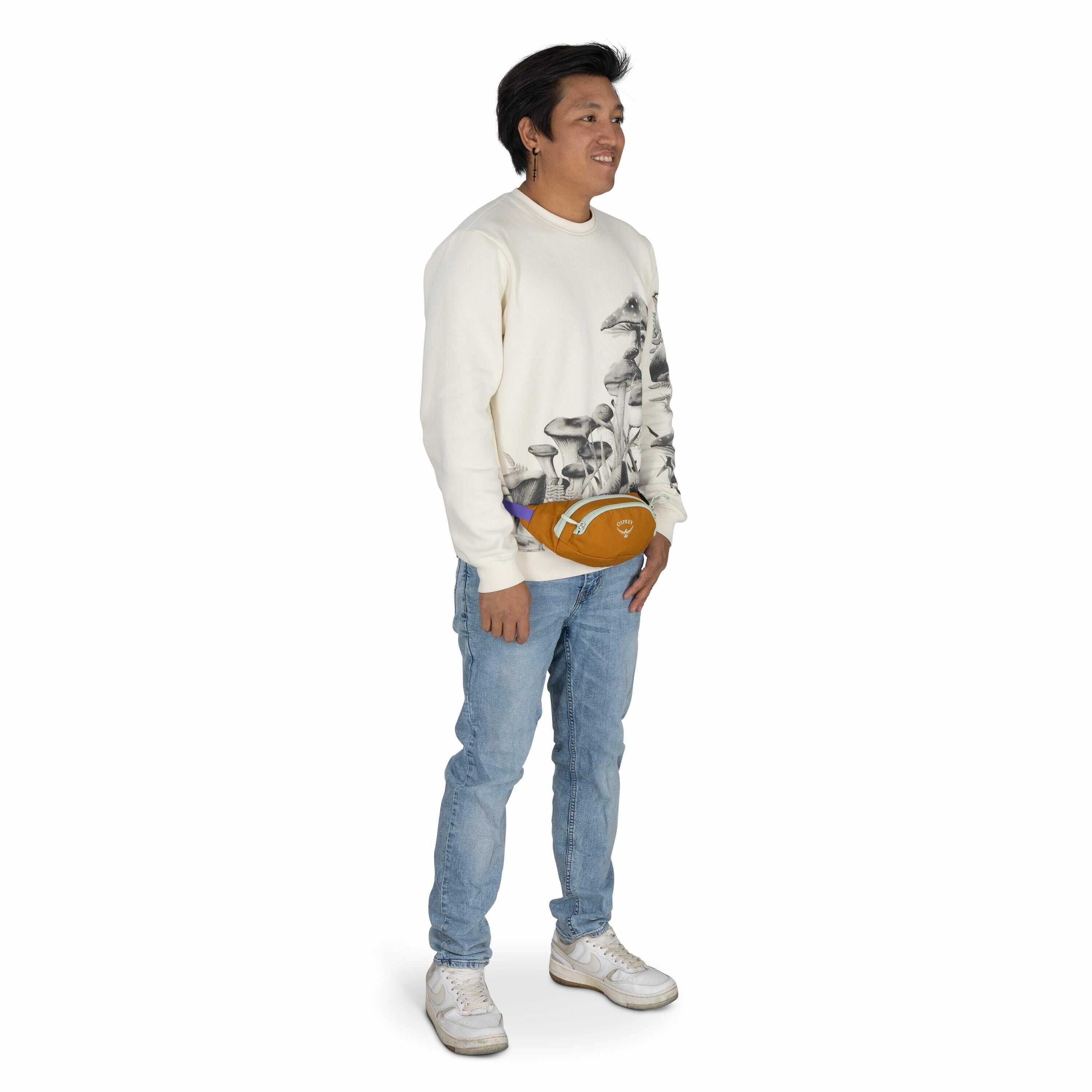 The image shows a person standing against a plain white background. They are wearing a light-colored sweatshirt with a detailed print of mushrooms on it, paired with light blue jeans. A mustard-colored fanny pack with purple accents is worn around the wai