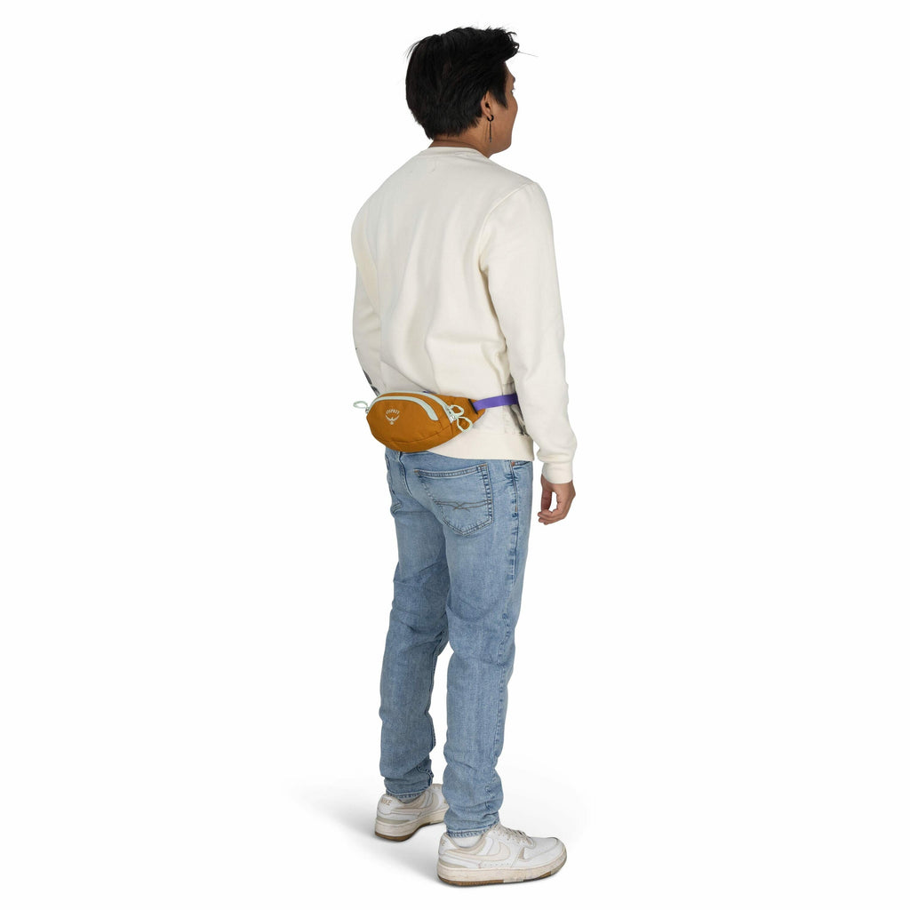 The image features a young man standing with his back turned towards the viewer. He is wearing a cream-colored long-sleeve shirt and light blue jeans, paired with white sneakers. A small orange waist bag with a purple strap is secured around his waist, an