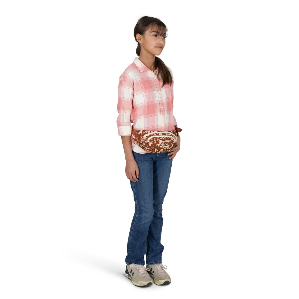 A person is standing against a plain white background wearing a casual outfit. They have on a pink-and-white plaid button-up shirt with rolled sleeves, blue jeans, and gray athletic shoes. A brown-and-cream patterned fanny pack is worn around the waist, t