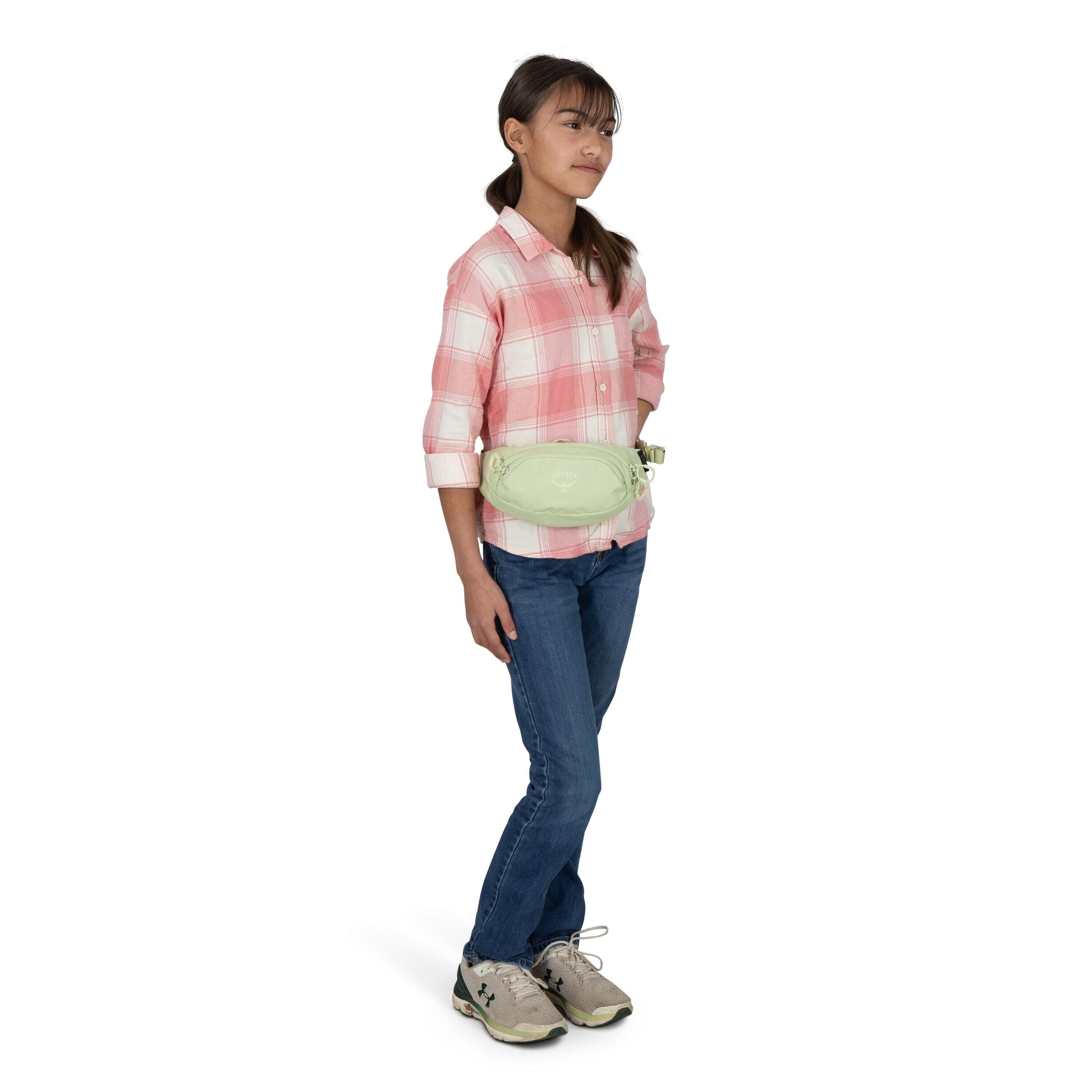 A person is standing against a white backdrop wearing a pink-and-white plaid button-up shirt, blue jeans, and gray sneakers. They have a light green Osprey waist pack worn around the hips. The individual's face is blurred, and their hands rest casually by