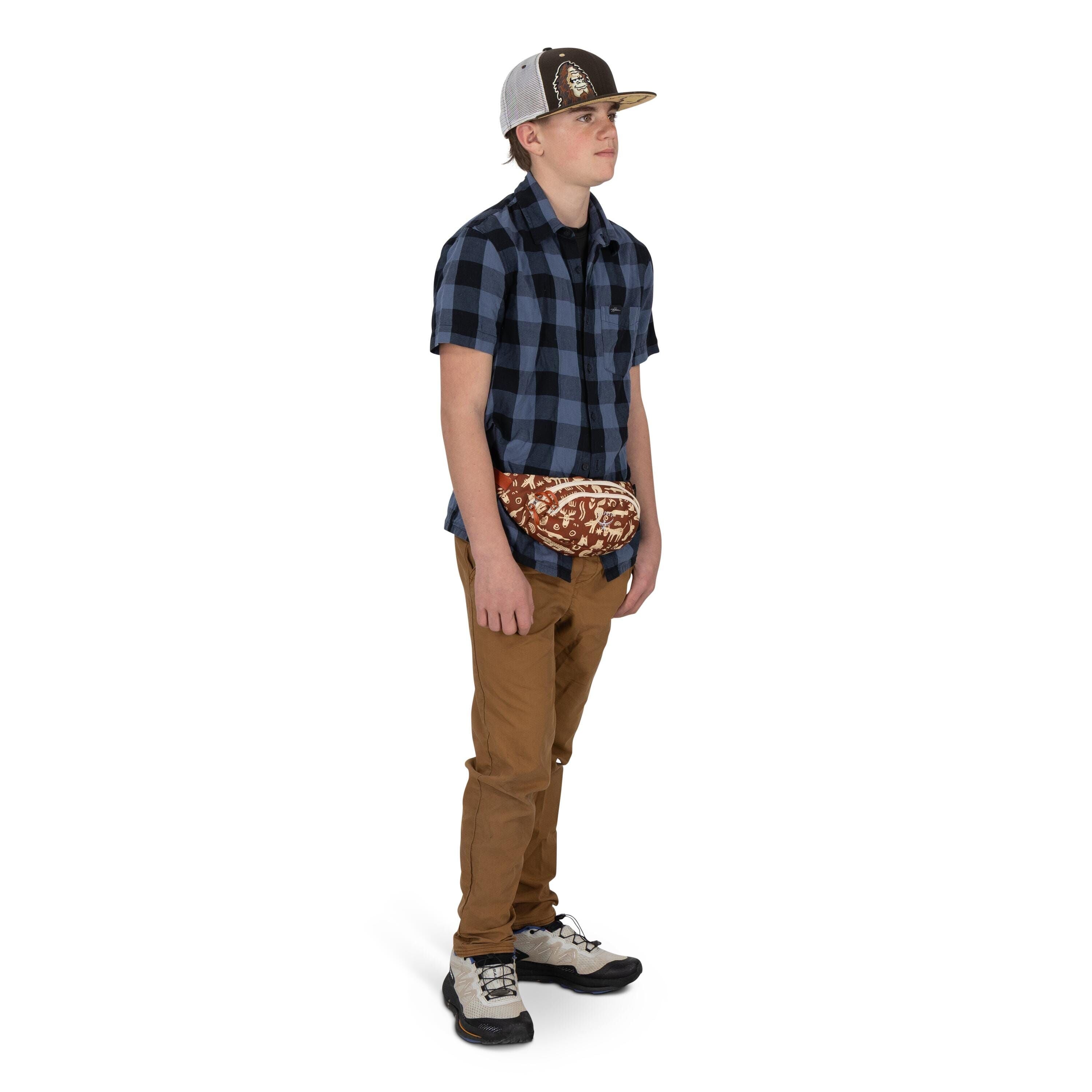 A young person stands against a white background wearing a blue-and-navy checkered short-sleeve shirt and tan pants. They have on a two-tone baseball cap and hiking-style sneakers. A patterned waist pack with a reddish-brown design is worn around the hips