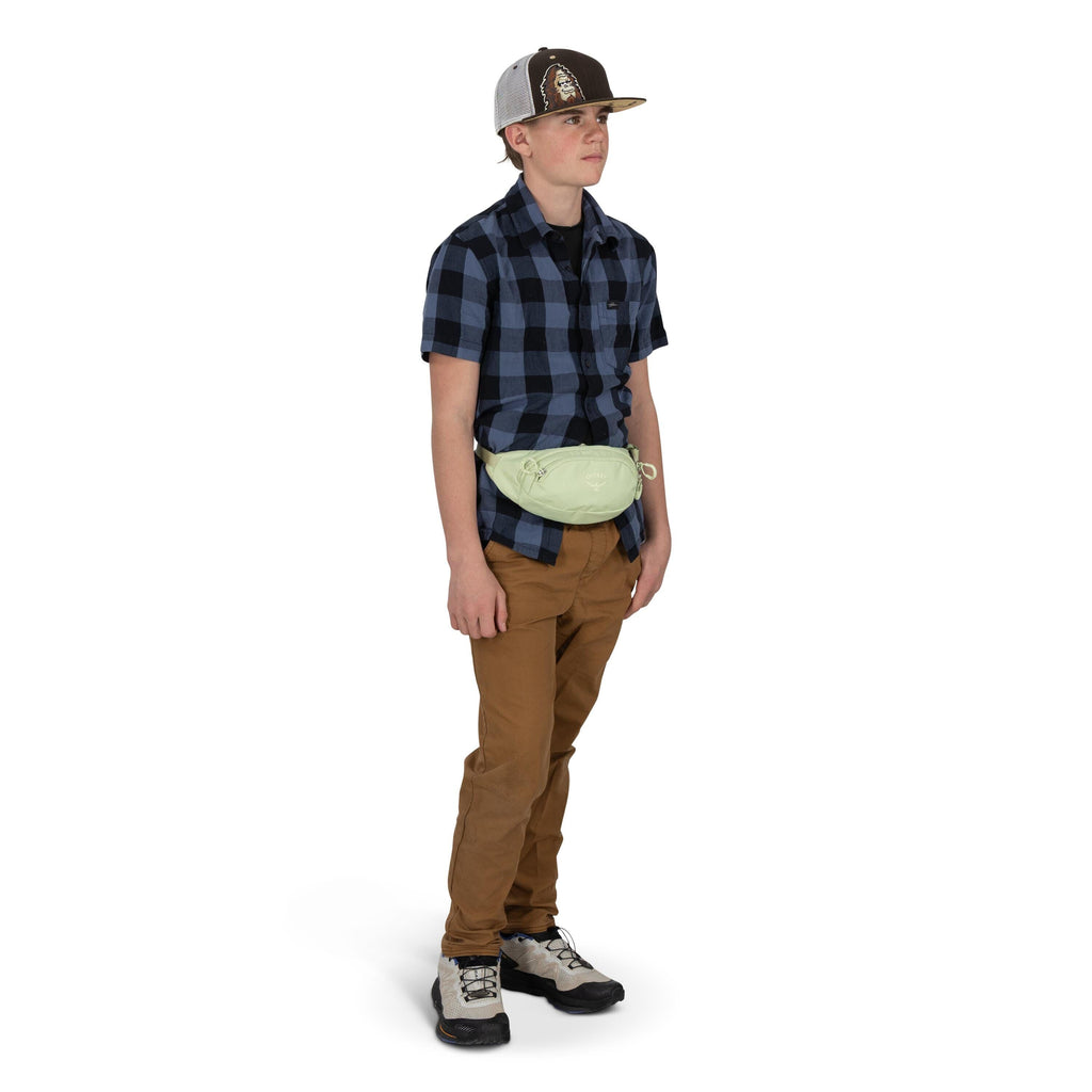 A person stands against a white background wearing a blue-and-black plaid short-sleeve shirt, tan pants, and hiking-style shoes. They have a light green waist pack strapped at the front and are resting their hands in their pockets. A two-tone baseball cap