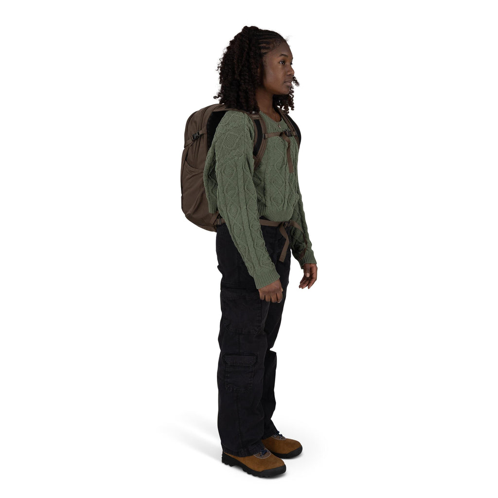 A person is standing in profile, wearing a green cable-knit sweater, black cargo pants, and brown hiking boots. They have a brown backpack with chest and waist straps clipped, suggesting they're prepared for outdoor activity. The individual has shoulder-l