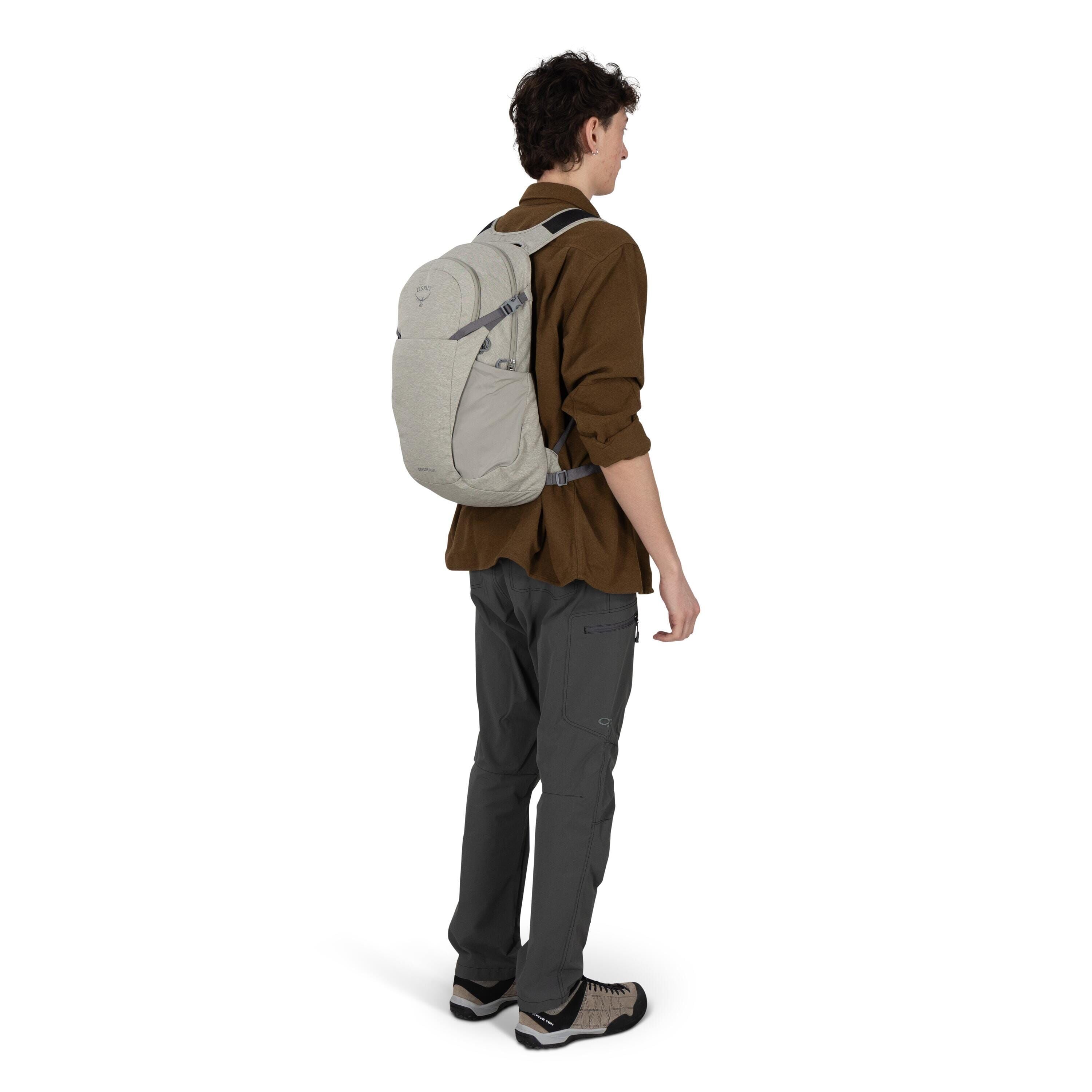A person is standing in a three-quarter rear view wearing an outdoor outfit with a light gray backpack. They have on a brown long-sleeve shirt, dark gray technical pants with a zip pocket on the thigh, and beige-and-black hiking shoes. The backpack appear