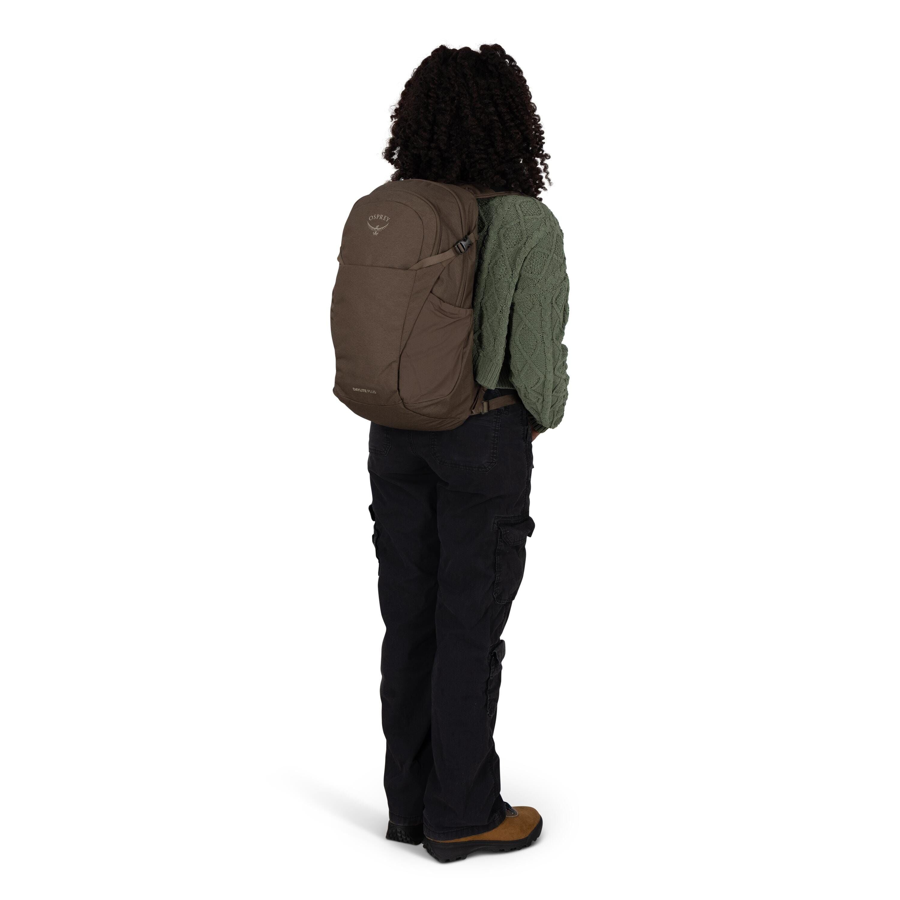 A person is standing with their back to the camera wearing a brown Osprey daypack. They have on a textured green sweater, black cargo pants, and brown hiking shoes. The scene is on a plain white background, emphasizing the fit and size of the backpack on