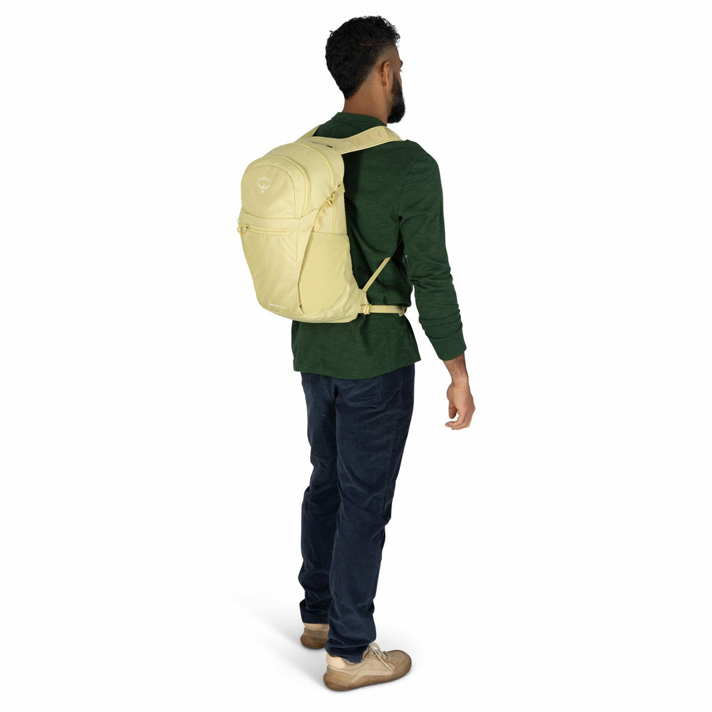 The image features a man standing and wearing a yellow backpack. He is facing slightly away from the camera, showing the back and side profile of the backpack. The man is dressed in a green long-sleeve shirt, dark blue pants, and brown shoes. The backpack