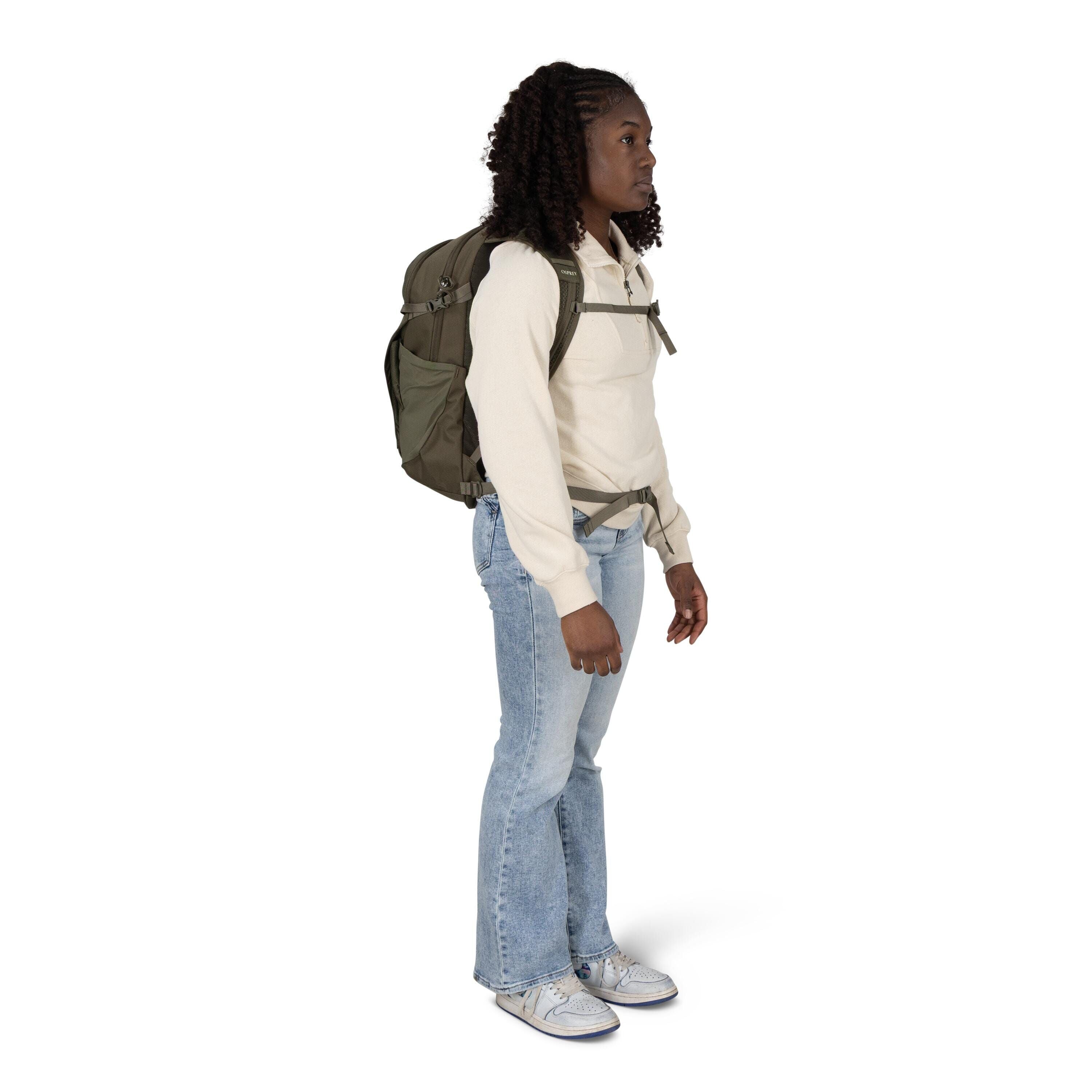 A person stands in a three-quarter profile wearing a light beige sweatshirt, light blue flared jeans, and gray-and-white sneakers. They have a large olive-green hiking backpack with chest and hip straps clipped across the torso. Their hands rest by their