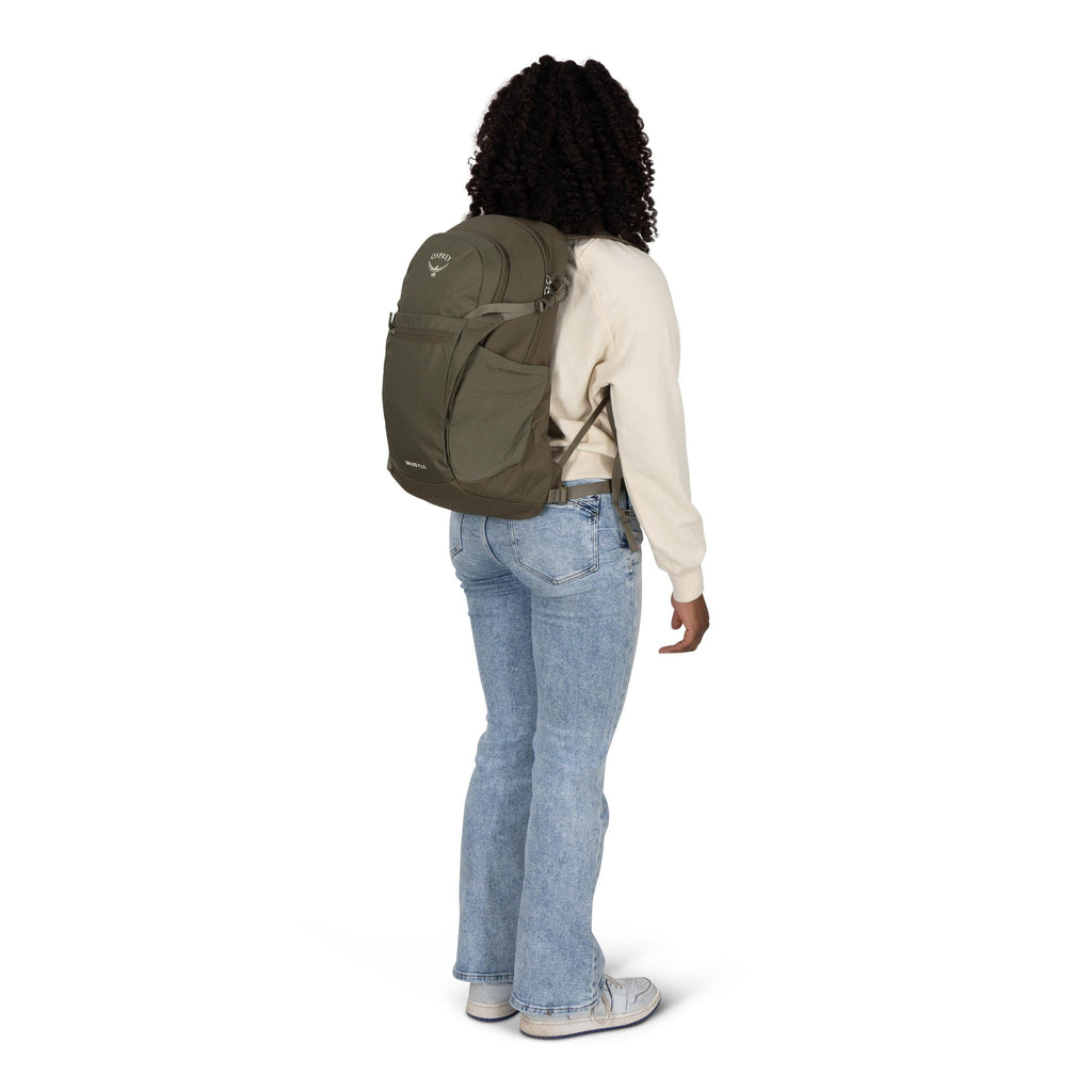 A person stands at a slight angle wearing a khaki green Osprey backpack. They are dressed casually in a cream sweatshirt, light blue jeans, and white sneakers. The backpack has multiple zippered compartments and side pockets, fitting snugly on their back.