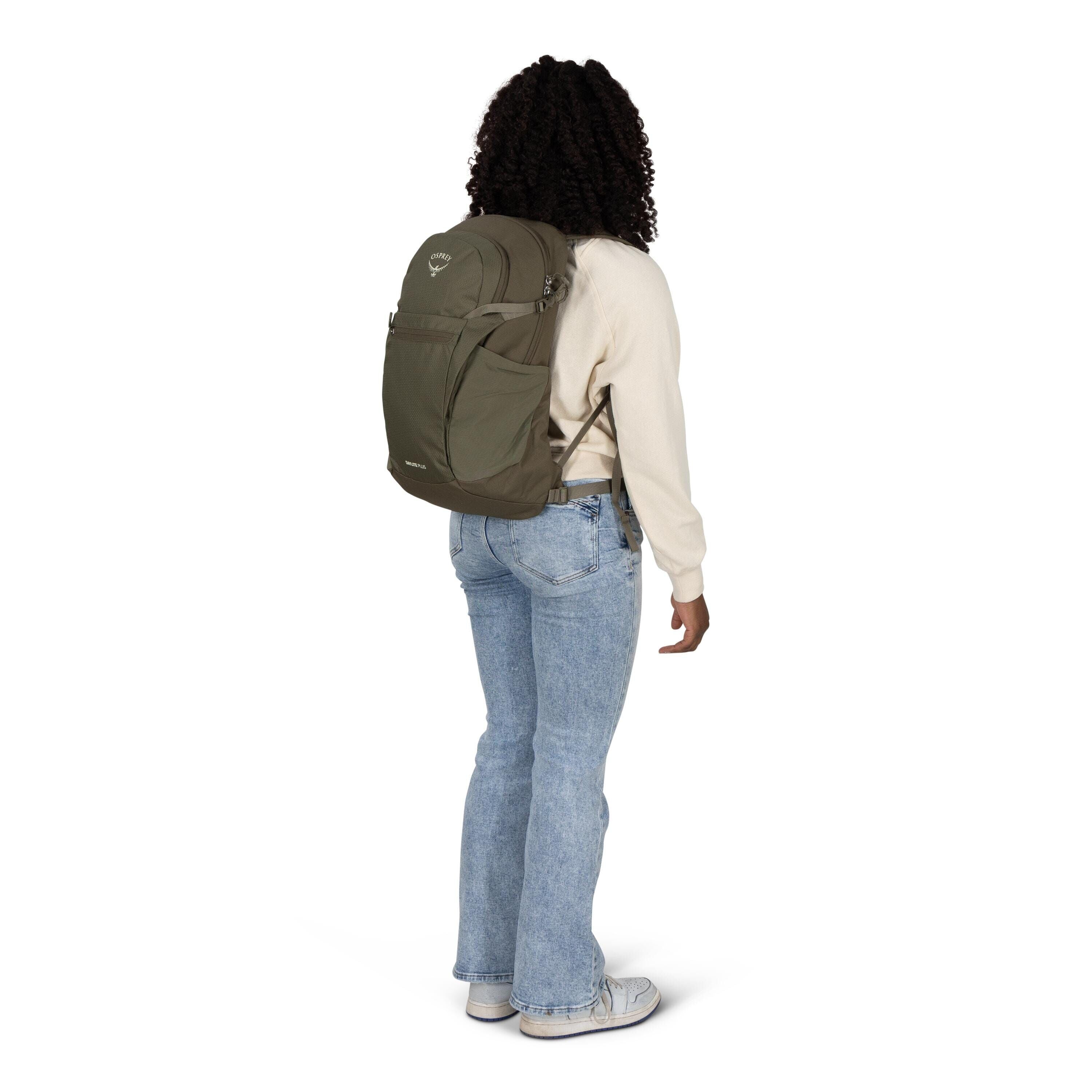 A person stands at a slight angle wearing a khaki green Osprey backpack. They are dressed casually in a cream sweatshirt, light blue jeans, and white sneakers. The backpack has multiple zippered compartments and side pockets, fitting snugly on their back.