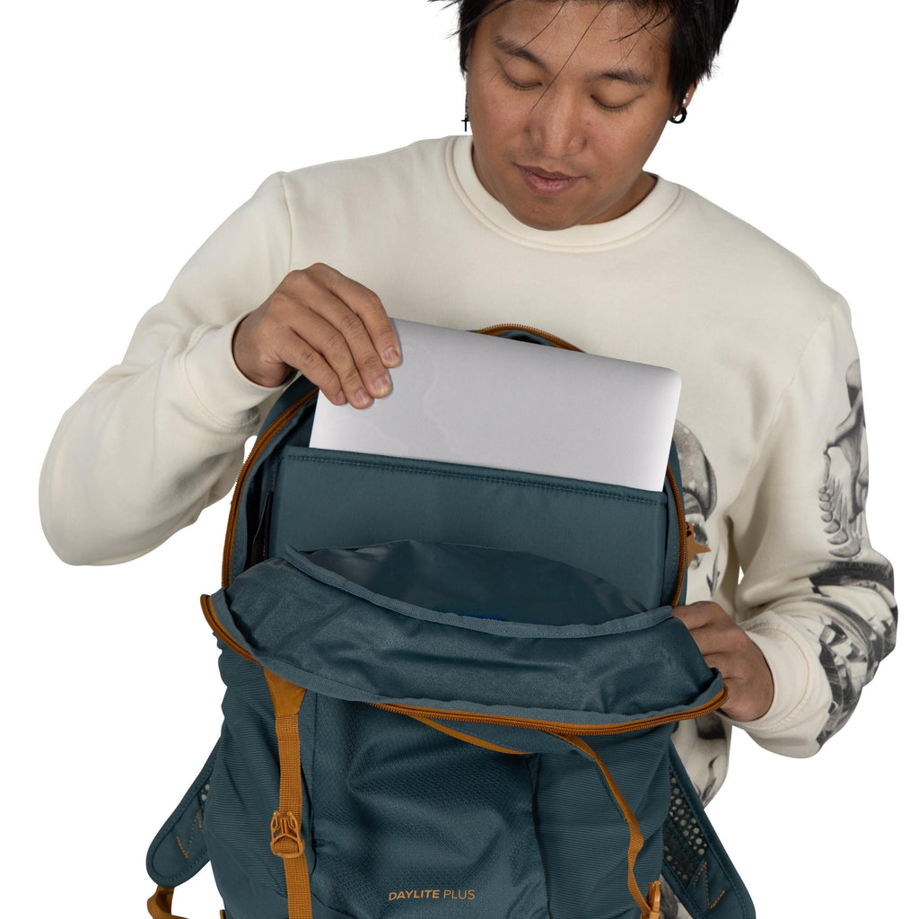 The image shows a person wearing a cream-colored sweatshirt with a black and white graphic design on the sleeves. The person is placing a silver laptop into a blue and orange Daylite Plus backpack. The backpack is open, revealing a compartment that appear