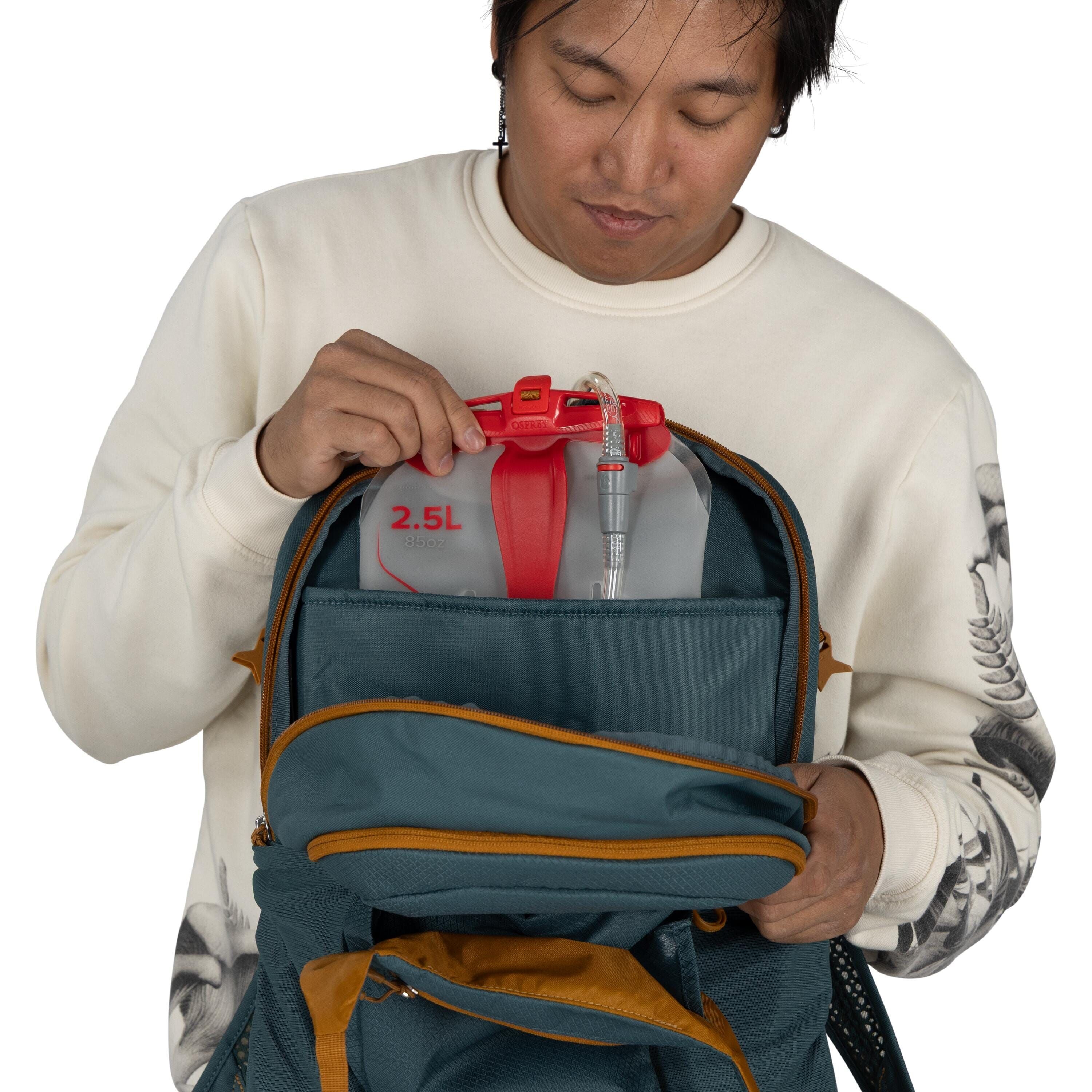 In the image, a person is holding a gray backpack with orange accents, which is partially open. Inside the backpack, the person is placing a red and transparent container labeled "2.5L" with a visible hose attached to it, suggesting it might be a water or