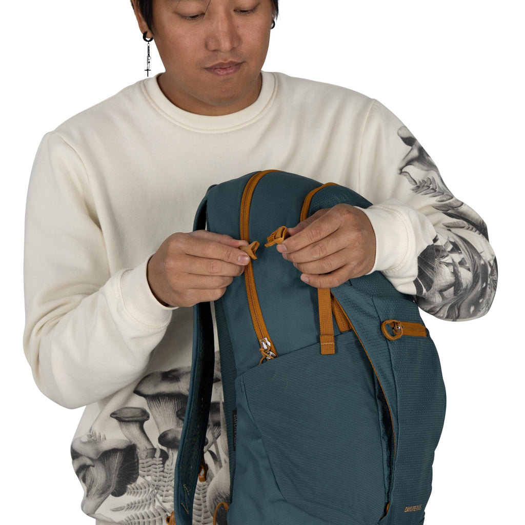The image features a person dressed in a cream-colored sweater adorned with a black and white mushroom design on the sleeves. They are holding a teal backpack, focusing on securing its two orange zippers. The person is wearing an earring shaped like a cro