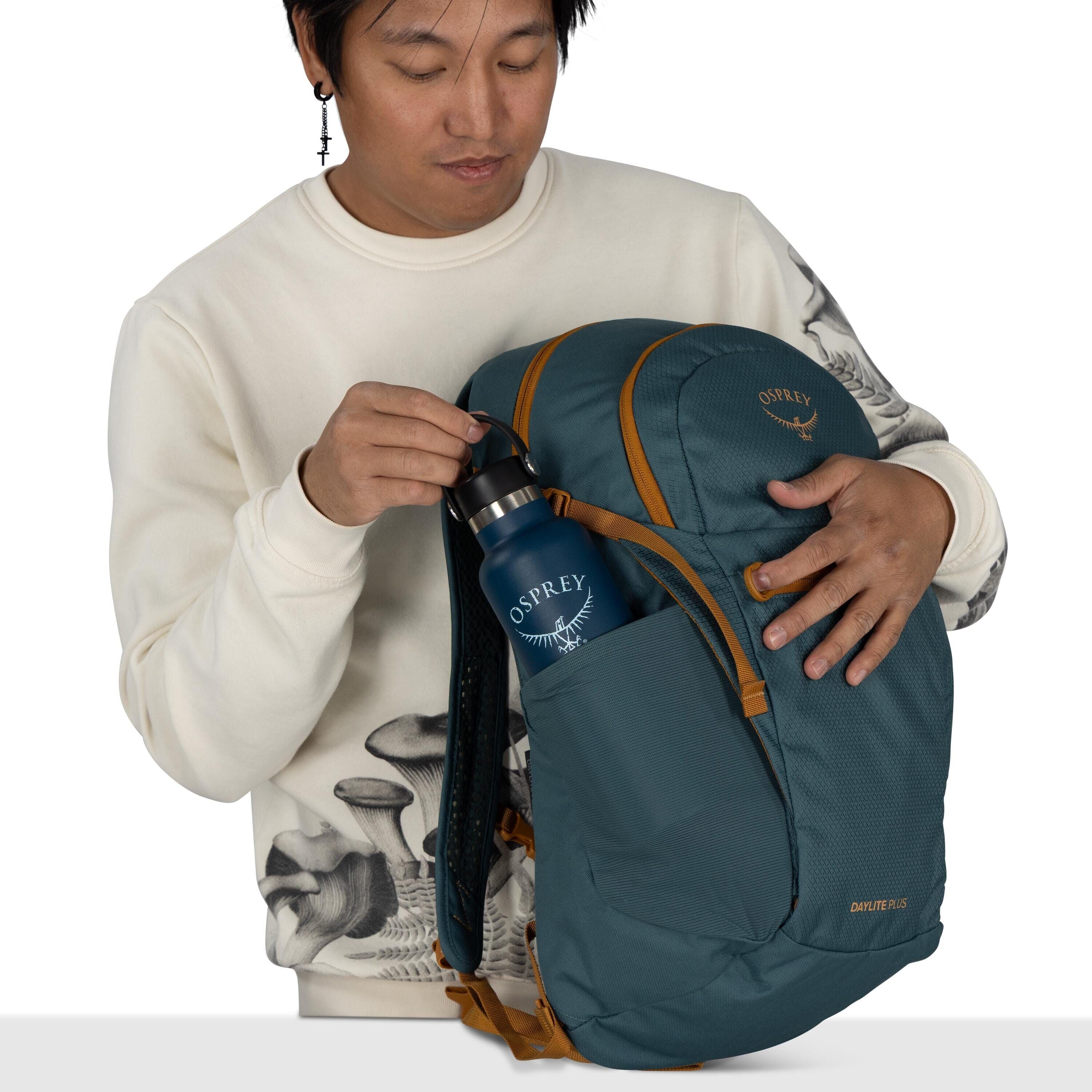 The image depicts a person holding a blue Osprey backpack in one hand while inserting a matching blue Osprey water bottle into a side pocket with the other hand. The backpack features orange accents and appears spacious, likely designed for outdoor or eve