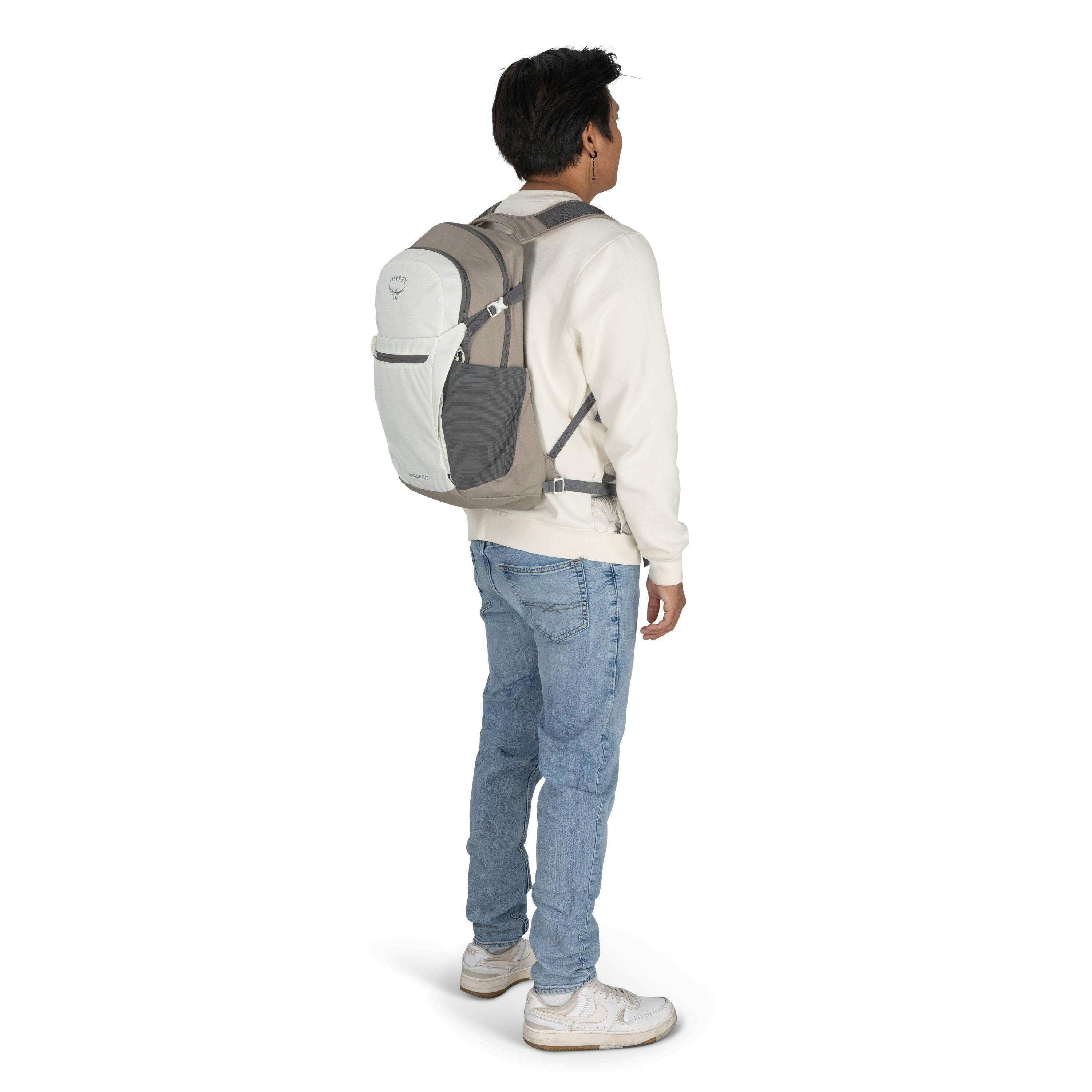 The image features a person standing and facing slightly away from the camera, wearing a gray and white Osprey backpack. The individual is dressed in a casual outfit consisting of a light-colored sweatshirt, blue jeans, and white sneakers. The backpack ap