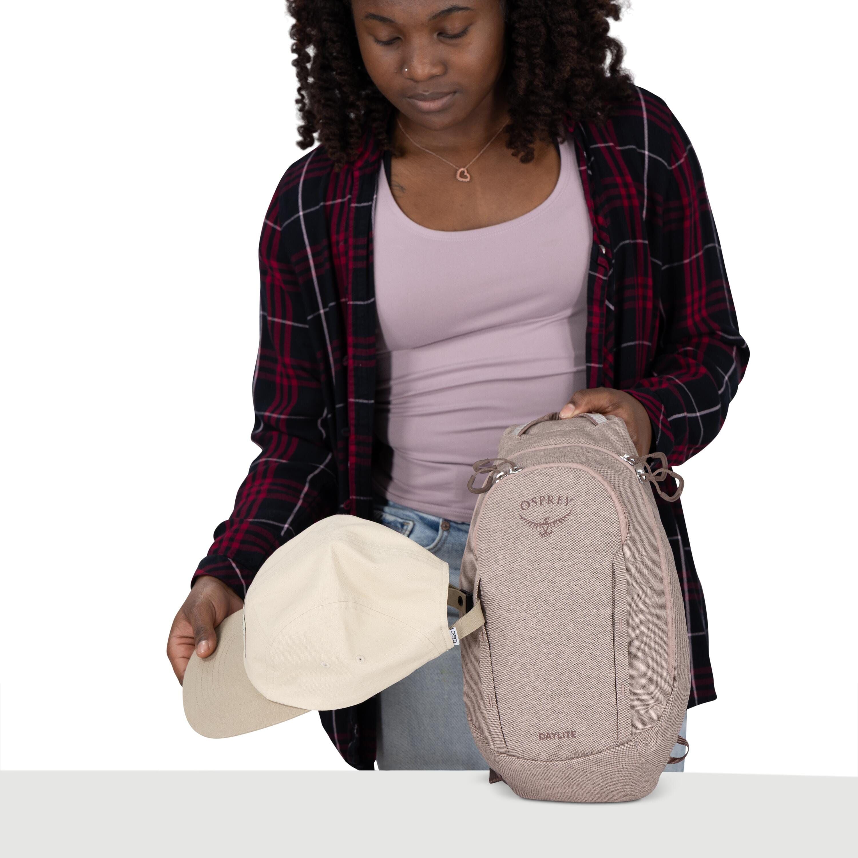 A person is holding a beige baseball cap and a small light-tan Osprey Daylite backpack. The backpack is upright with visible zippers and the Osprey logo on the front. The person wears a lavender tank top under a black-and-red plaid shirt and light denim j