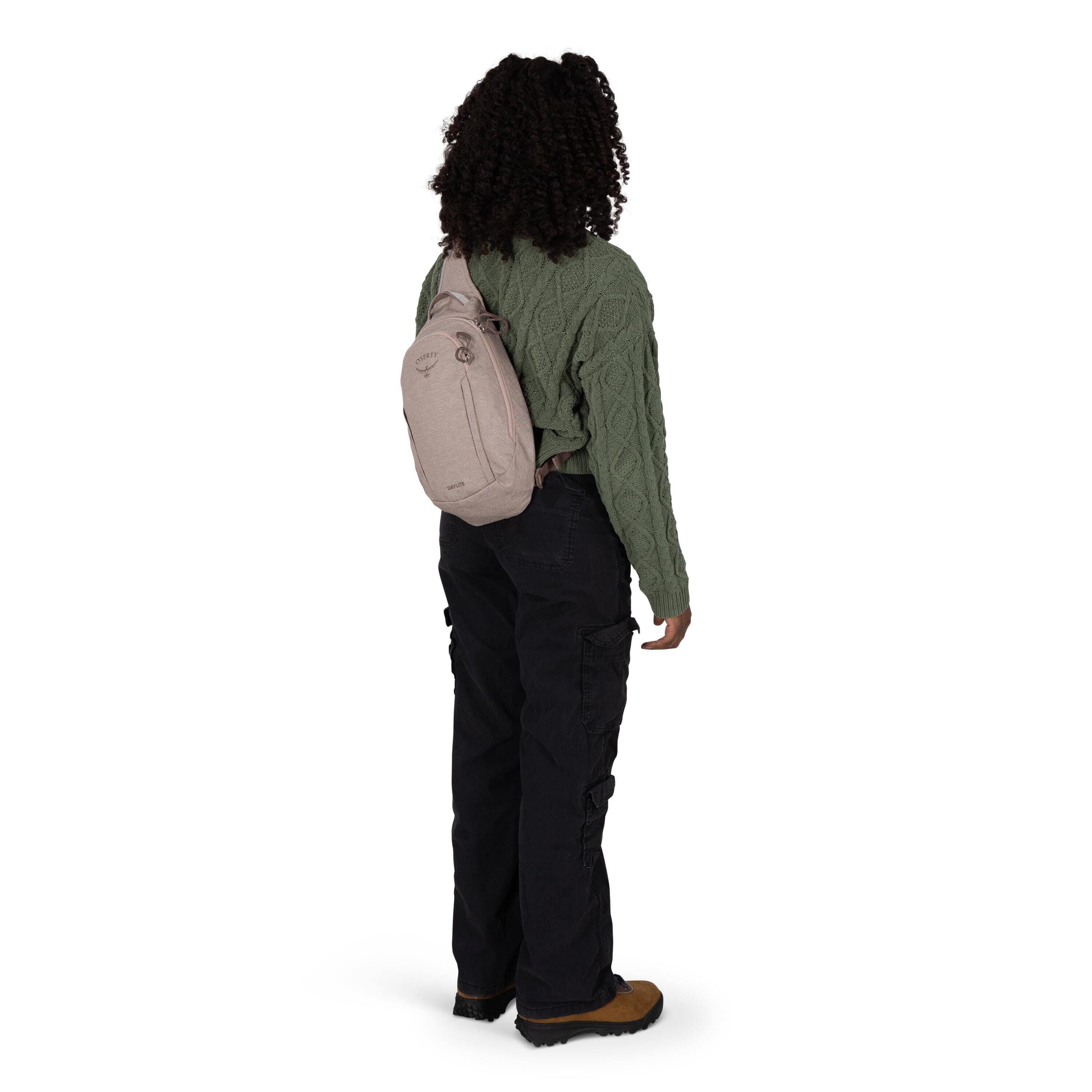 A person stands facing slightly away, wearing a small beige sling backpack over one shoulder. They have on a textured green cable-knit sweater, loose black cargo pants, and brown hiking-style shoes. The scene is on a plain white background, highlighting t