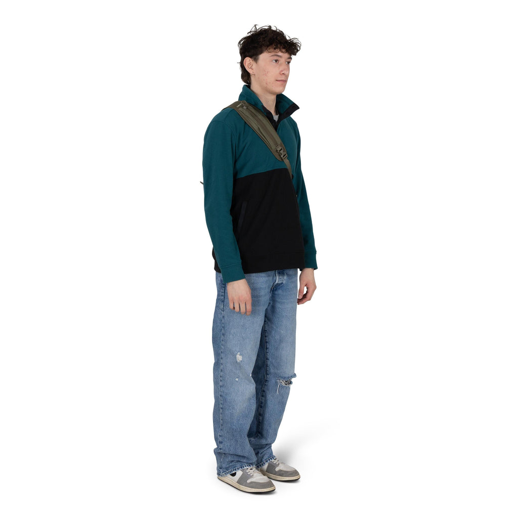 A person stands against a white background wearing a casual outfit. They have on a teal and black quarter-zip pullover, light blue baggy jeans with distressed patches, and gray sneakers. An olive-green crossbody sling bag is worn across the chest. The pos