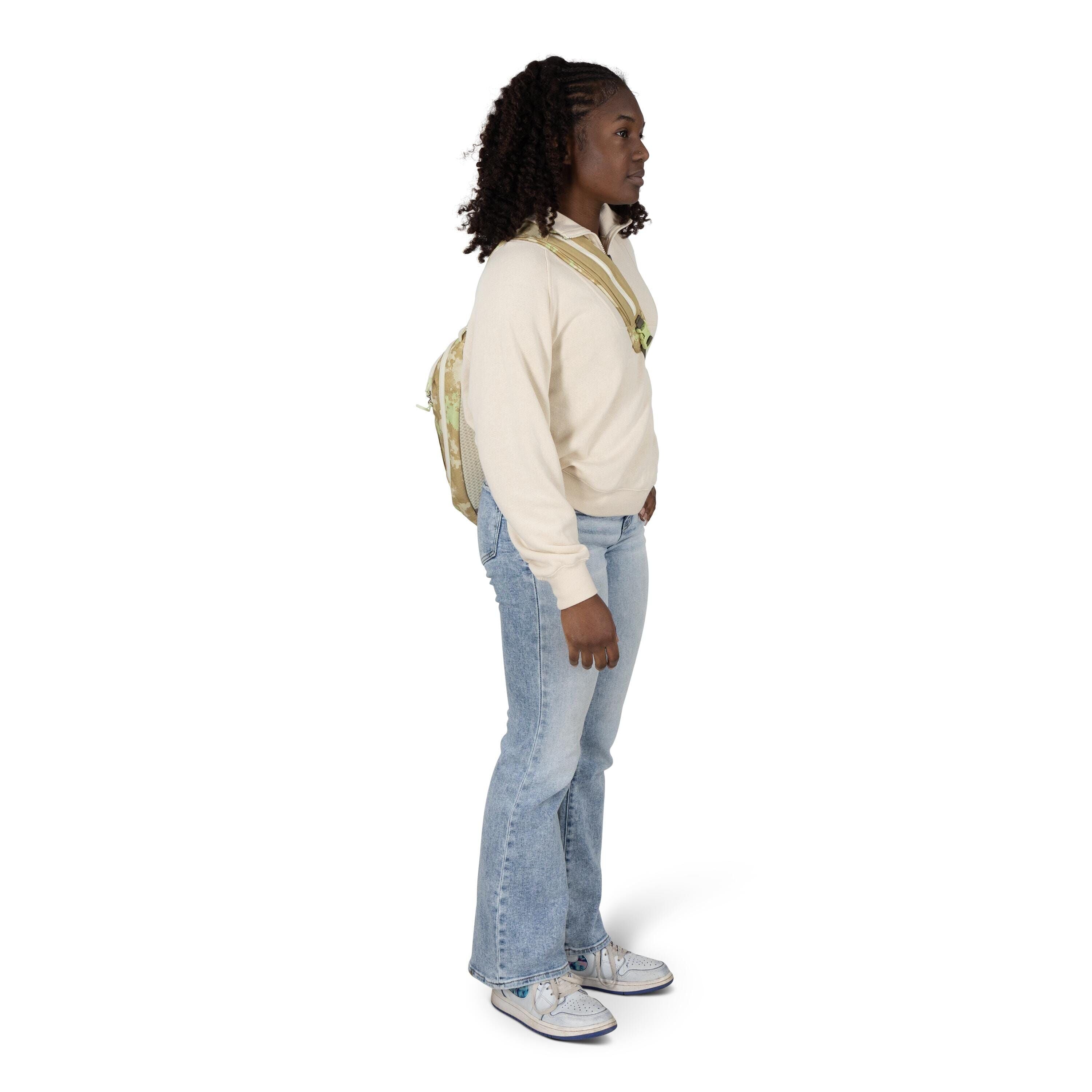 A person stands in a side profile pose against a white background, wearing a cream-colored pullover, light blue bootcut jeans, and patterned sneakers. They have curly shoulder-length hair and carry a light-colored backpack with floral designs. One hand re
